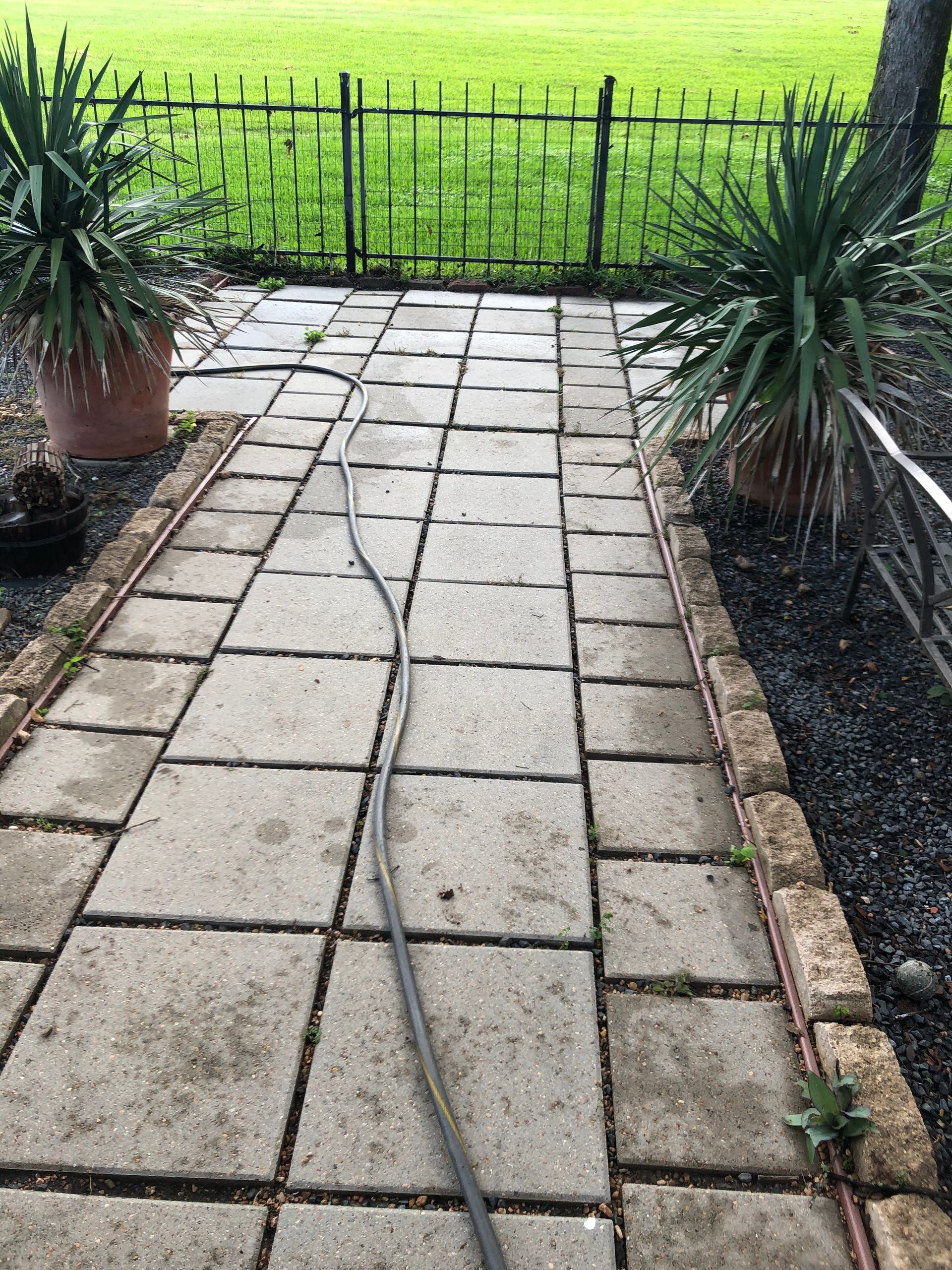 Stone garden path with a loose cable running down the center, bordered by potted plants and a fence.