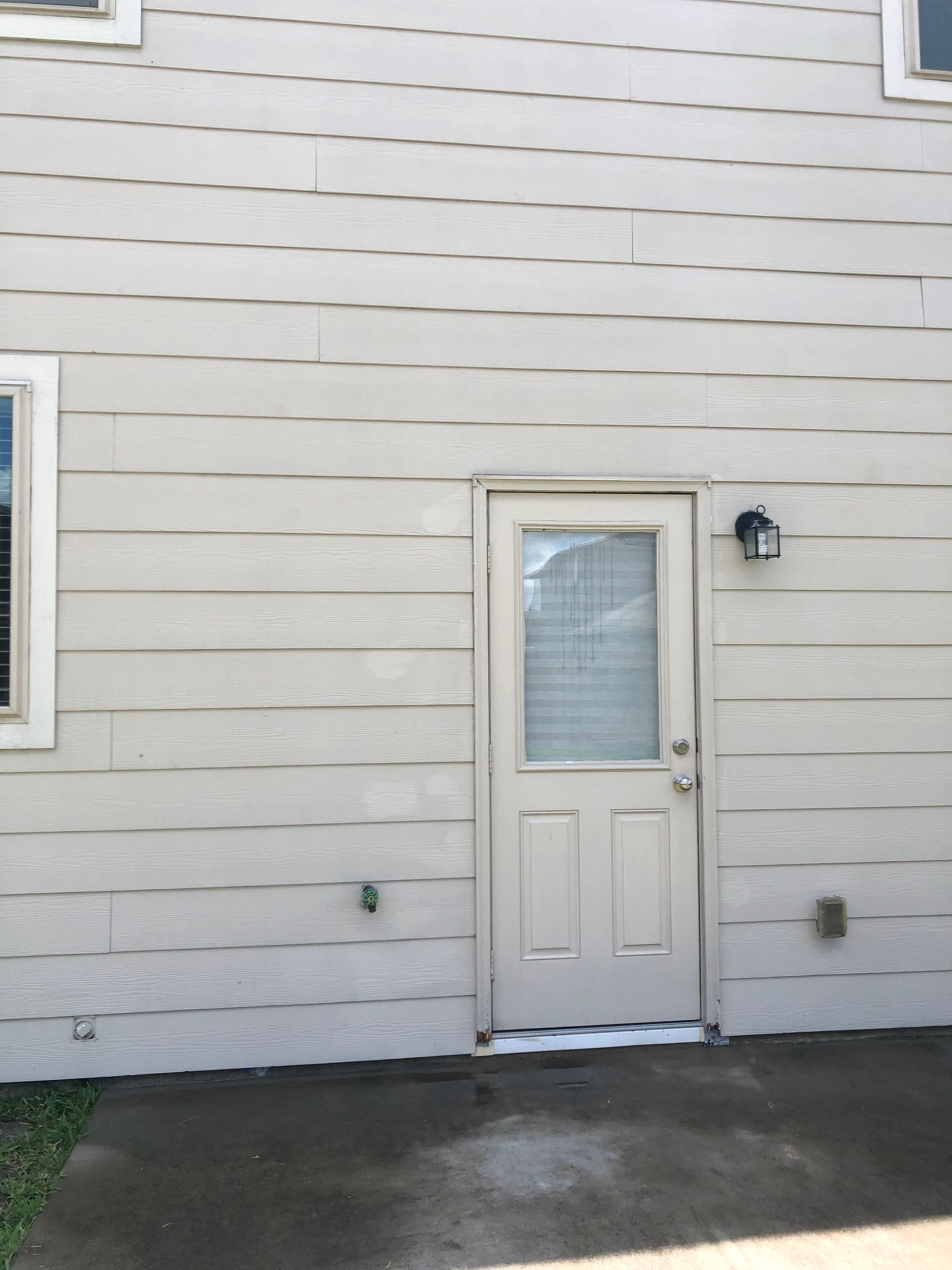 White house exterior with a small door, window, and wall-mounted light beside a concrete patio.