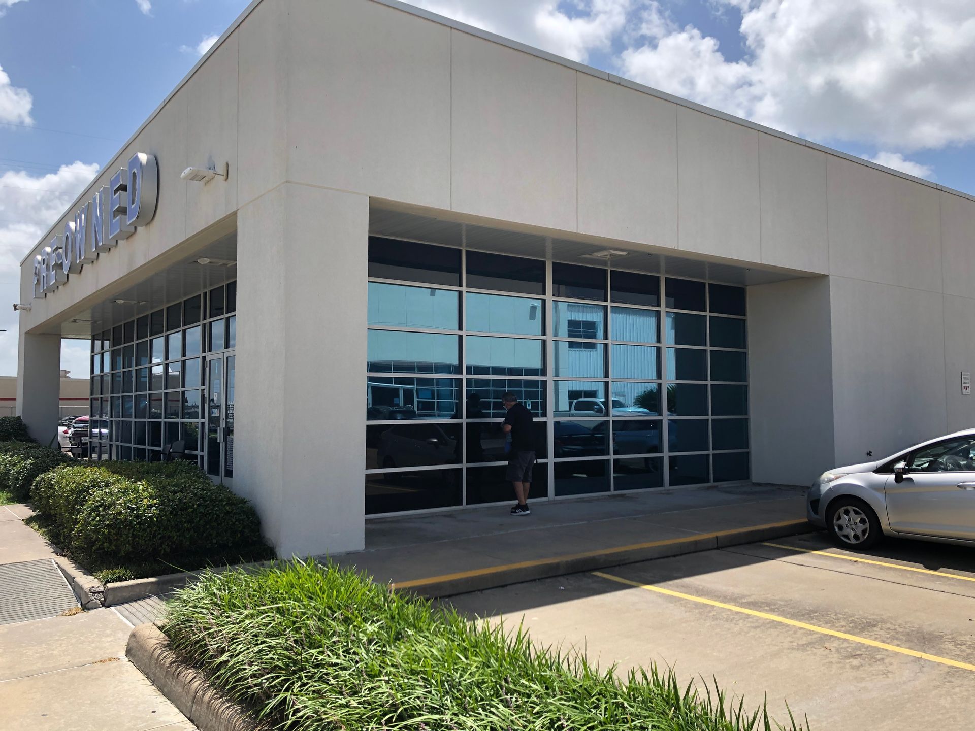 Car dealership storefront with large glass windows, white exterior, and a parked car in front