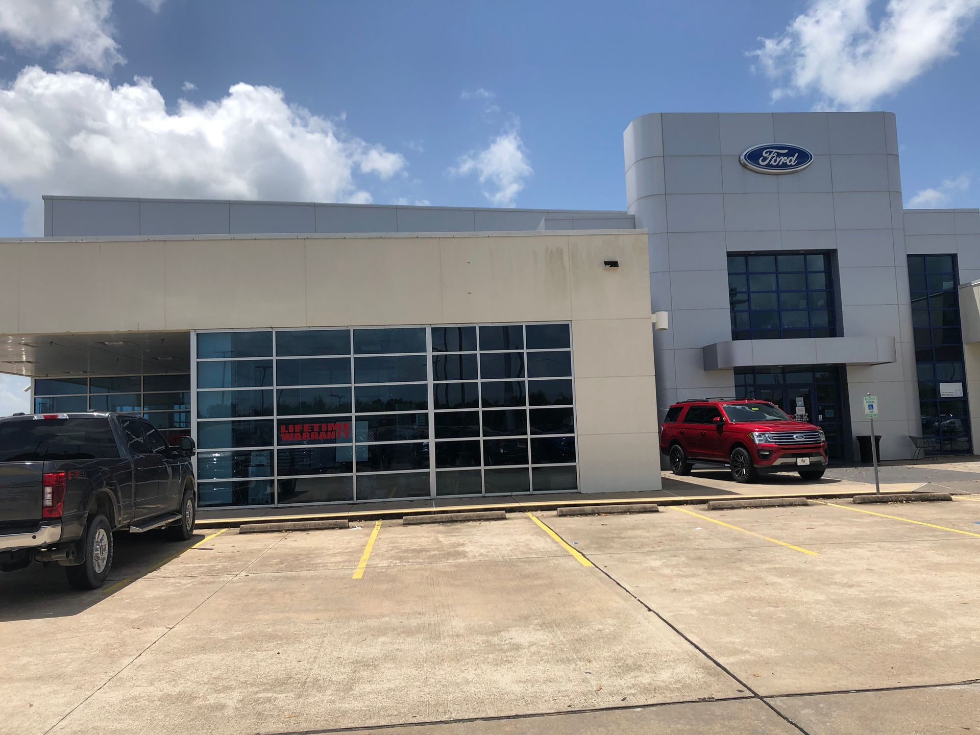 Ford dealership exterior with glass service bays, a red SUV, and a black truck under a cloudy sky