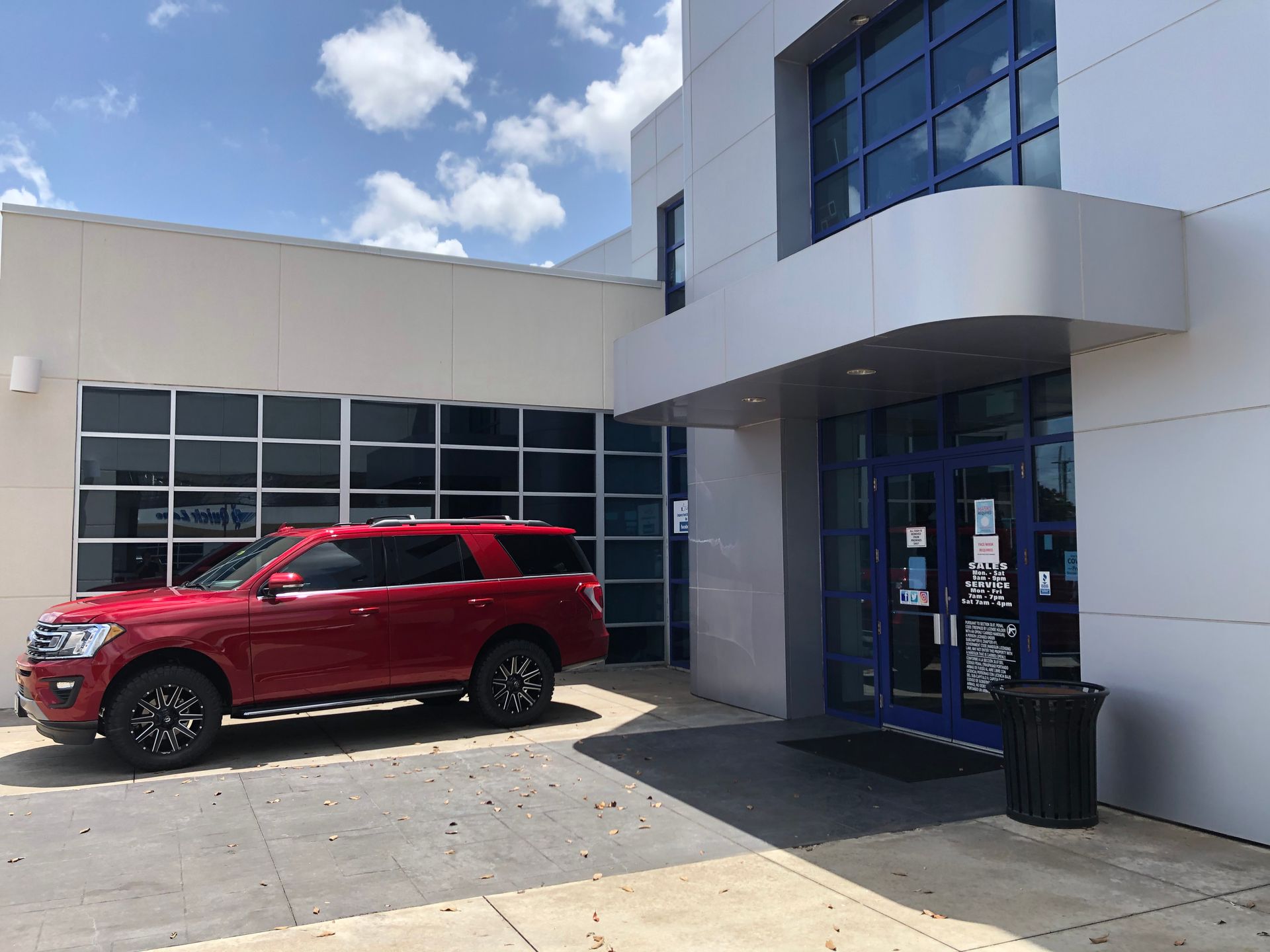 Red SUV parked outside a modern building entrance with glass windows and a trash can nearby