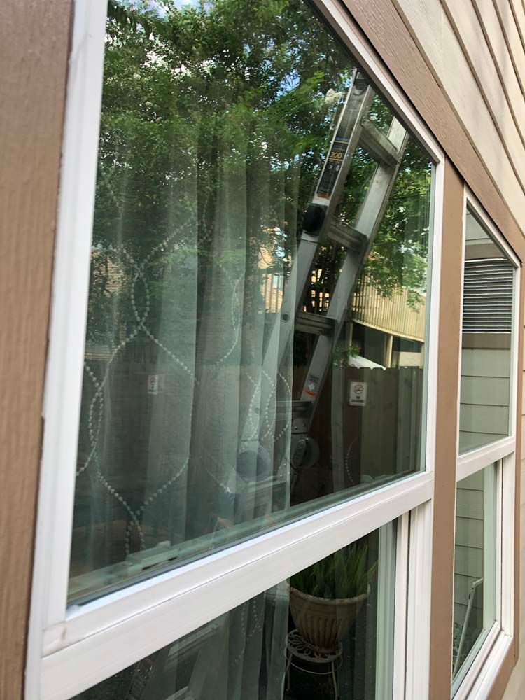 Open window reflecting a ladder, with curtains visible inside and house siding outside