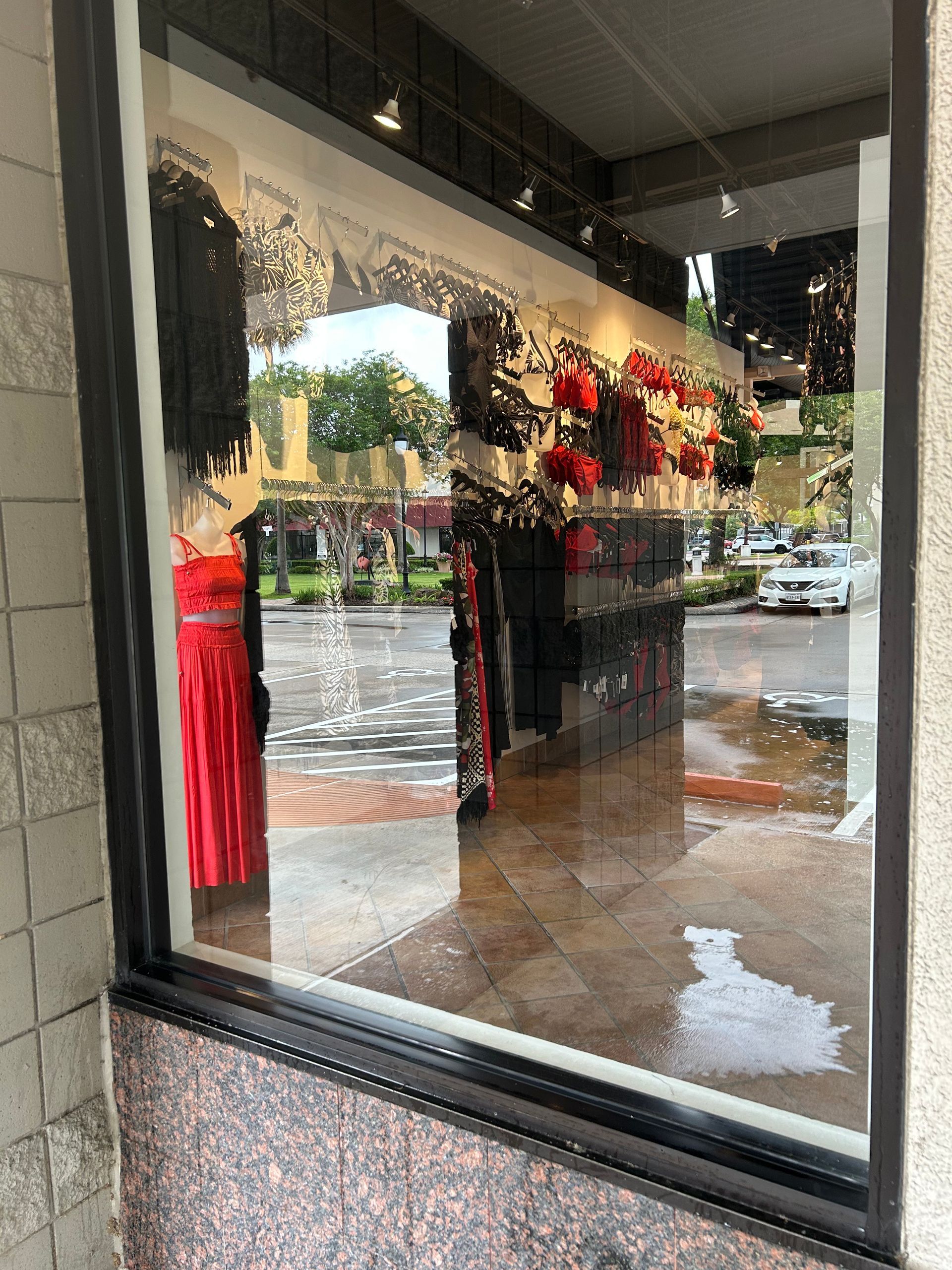 Clothing store window display with red dress, black garments, and floral decorations reflected on wet pavement