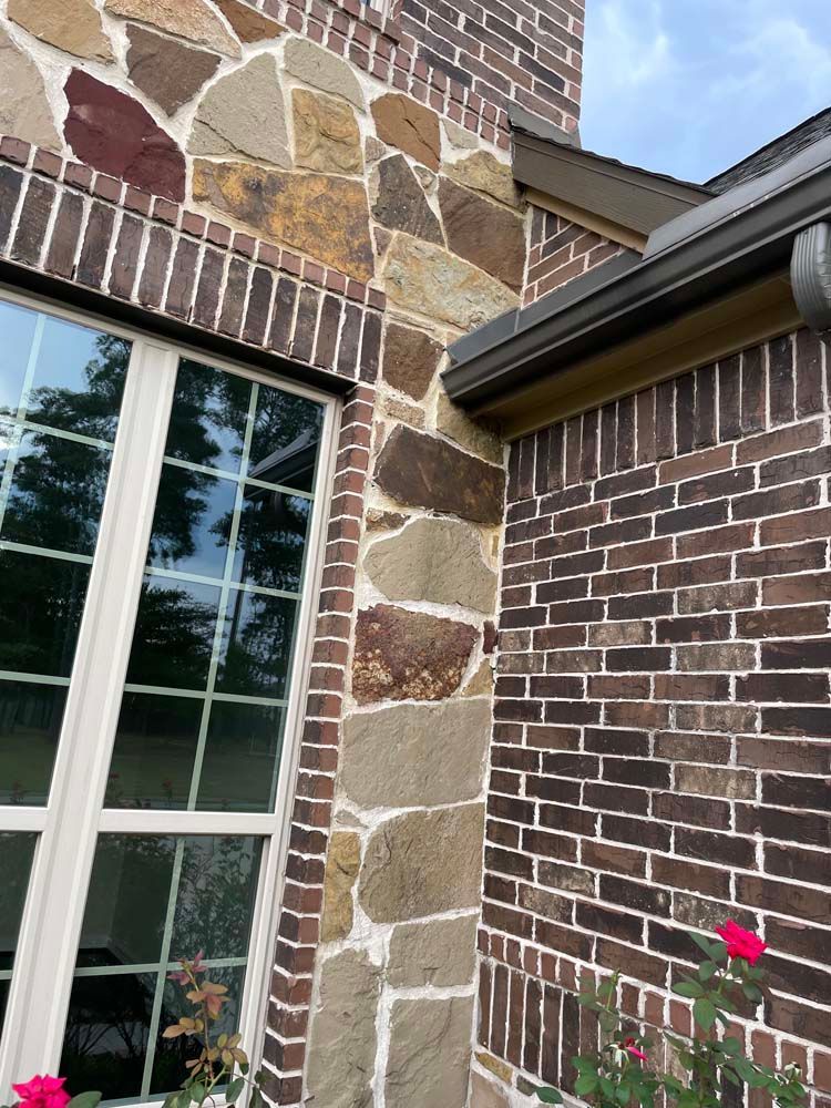 Brick and stone house exterior with a window, downspout, and a small flower bed.
