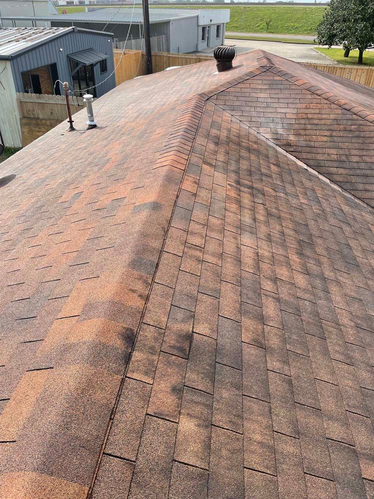 Weathered shingle roof with a central ridge vent beside a barn-like building and open fields.