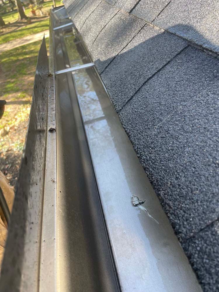 Metal gutter running along a shingled roof edge beside trees and a house wall