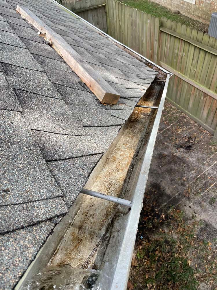 Damaged roof gutter filled with debris beside gray shingles and a wooden fence