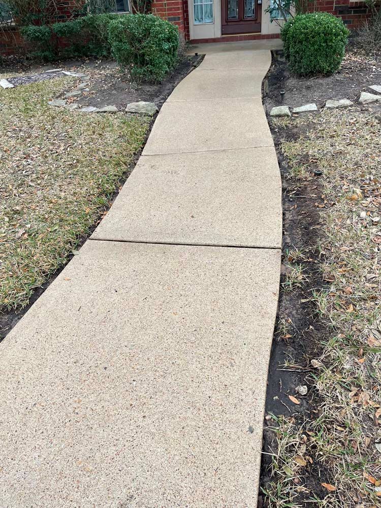 Concrete walkway leading to a front door, bordered by sparse grass and shrubs.