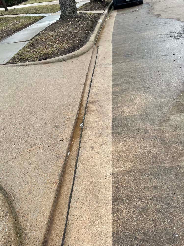 Cracked concrete sidewalk with a curb and drainage gap beside a wet street and parked car.