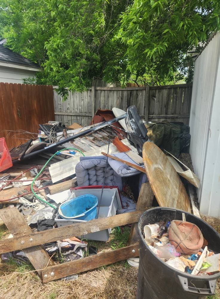Backyard piled with broken furniture, boards, buckets, and trash near a wooden fence and trees