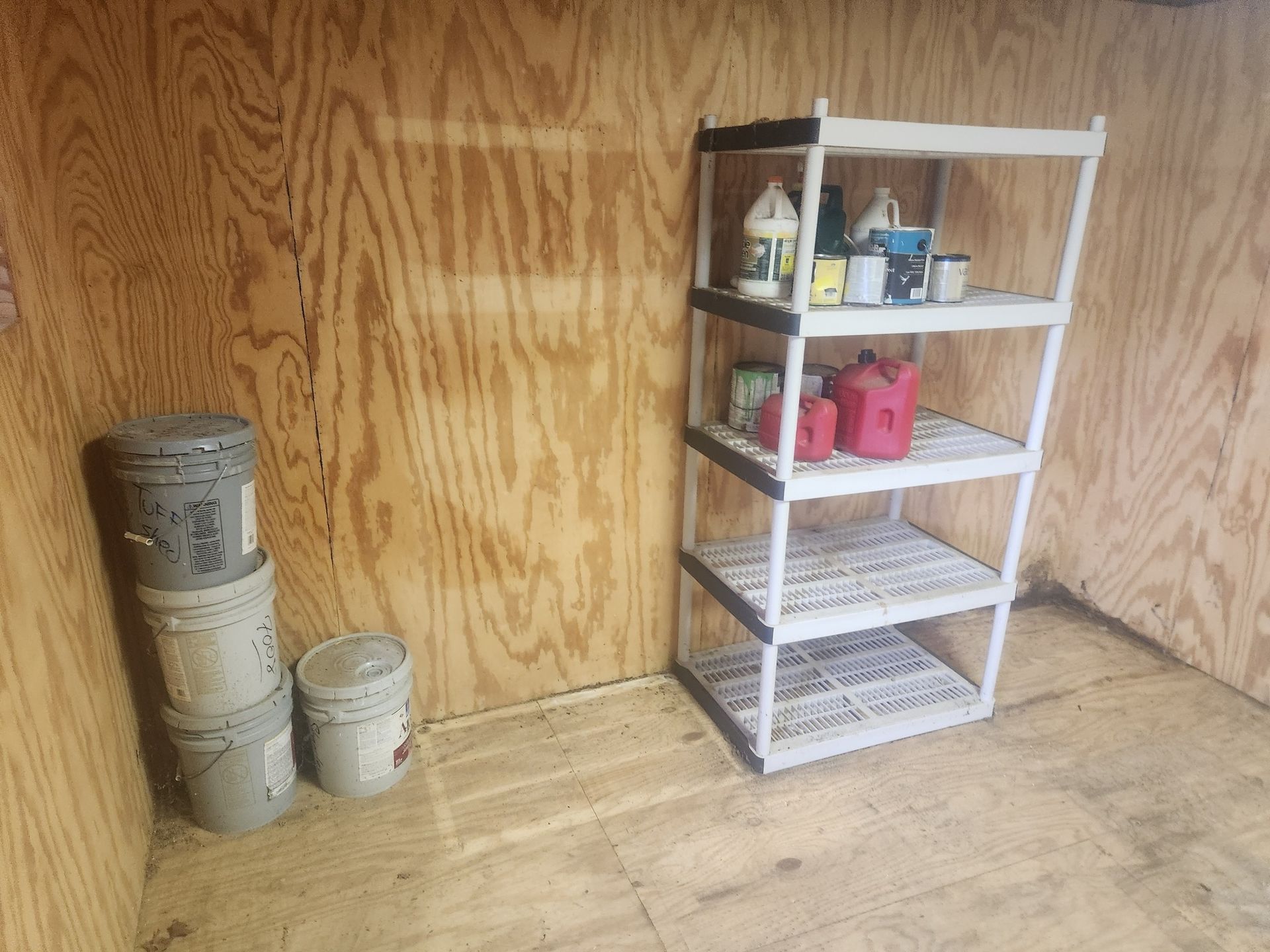Small plywood room with a white shelving unit holding tools and pink containers, plus stacked buckets on the left.