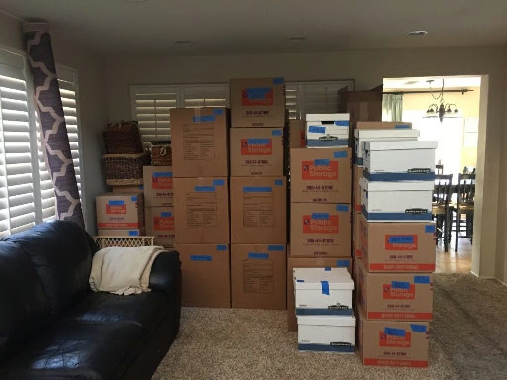 Living room filled with stacked moving boxes beside a black leather couch and window blinds