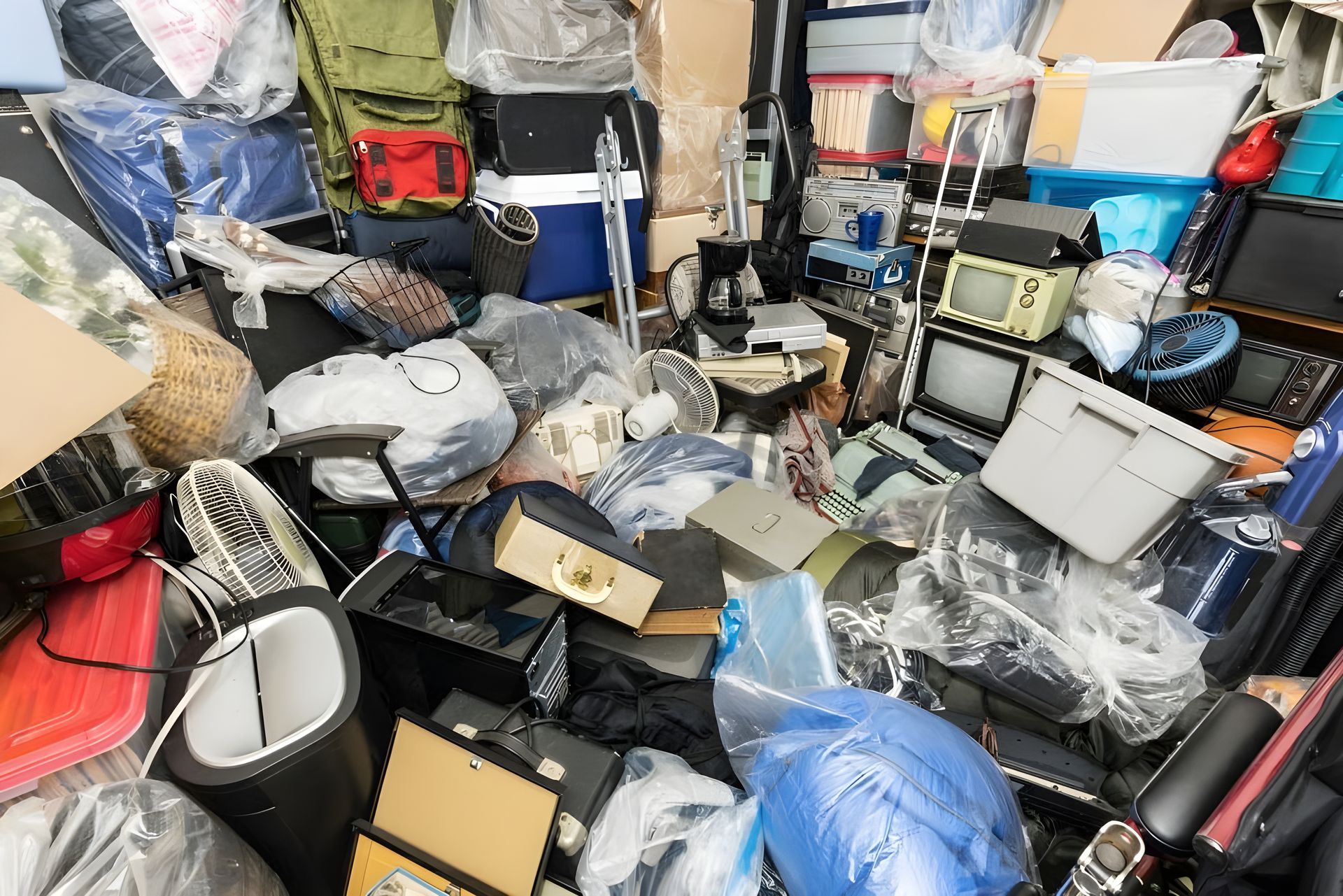 Cluttered storage room filled with stacked boxes, bags, and miscellaneous electronics and household items.