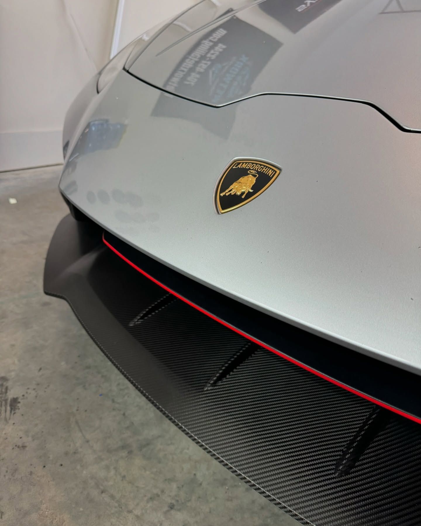Silver Lamborghini front with carbon fiber spoiler, red accent, and gold emblem.