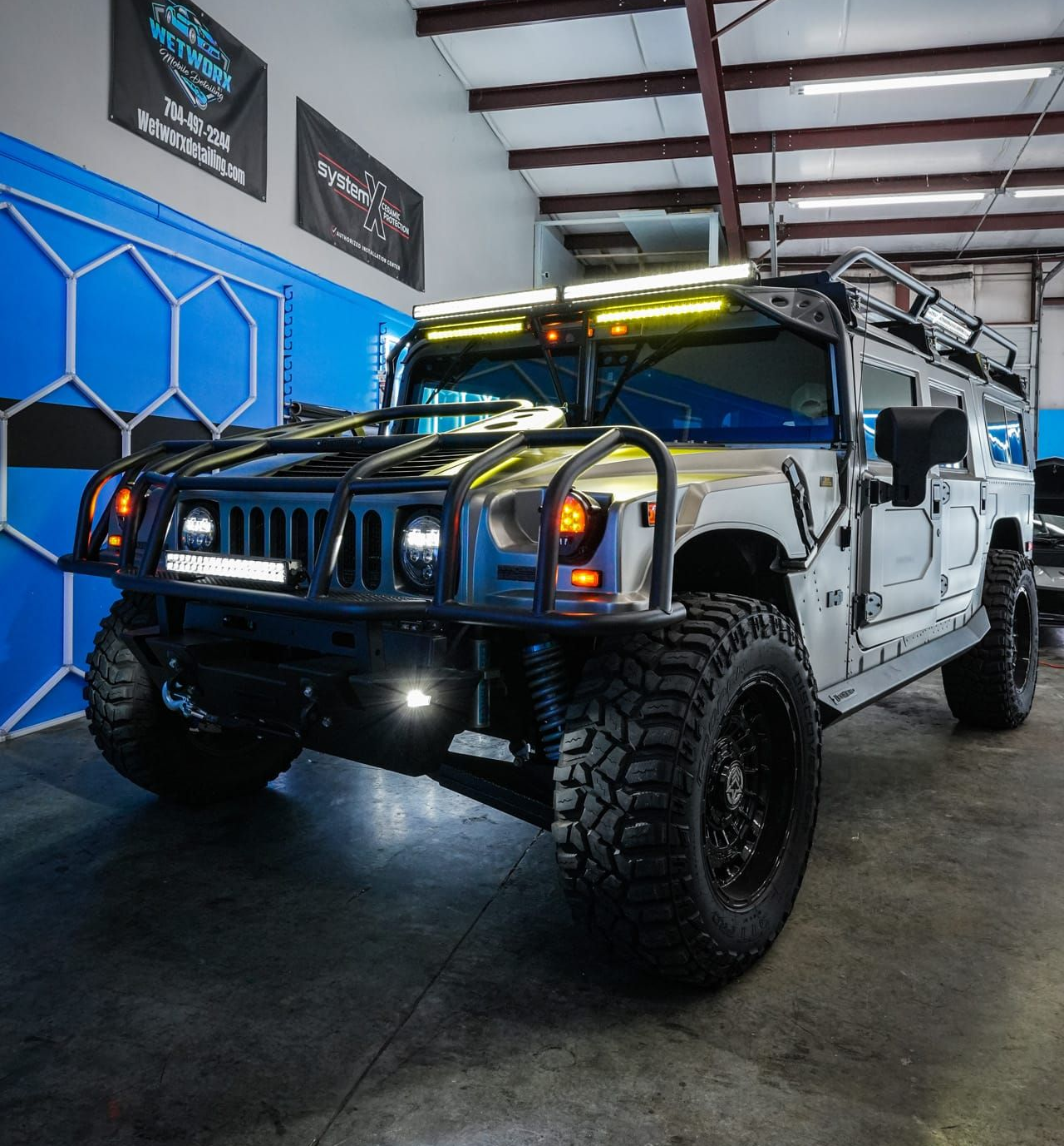 Modified Hummer H2 in a shop, with a roof rack, black wheels, and LED light bars.