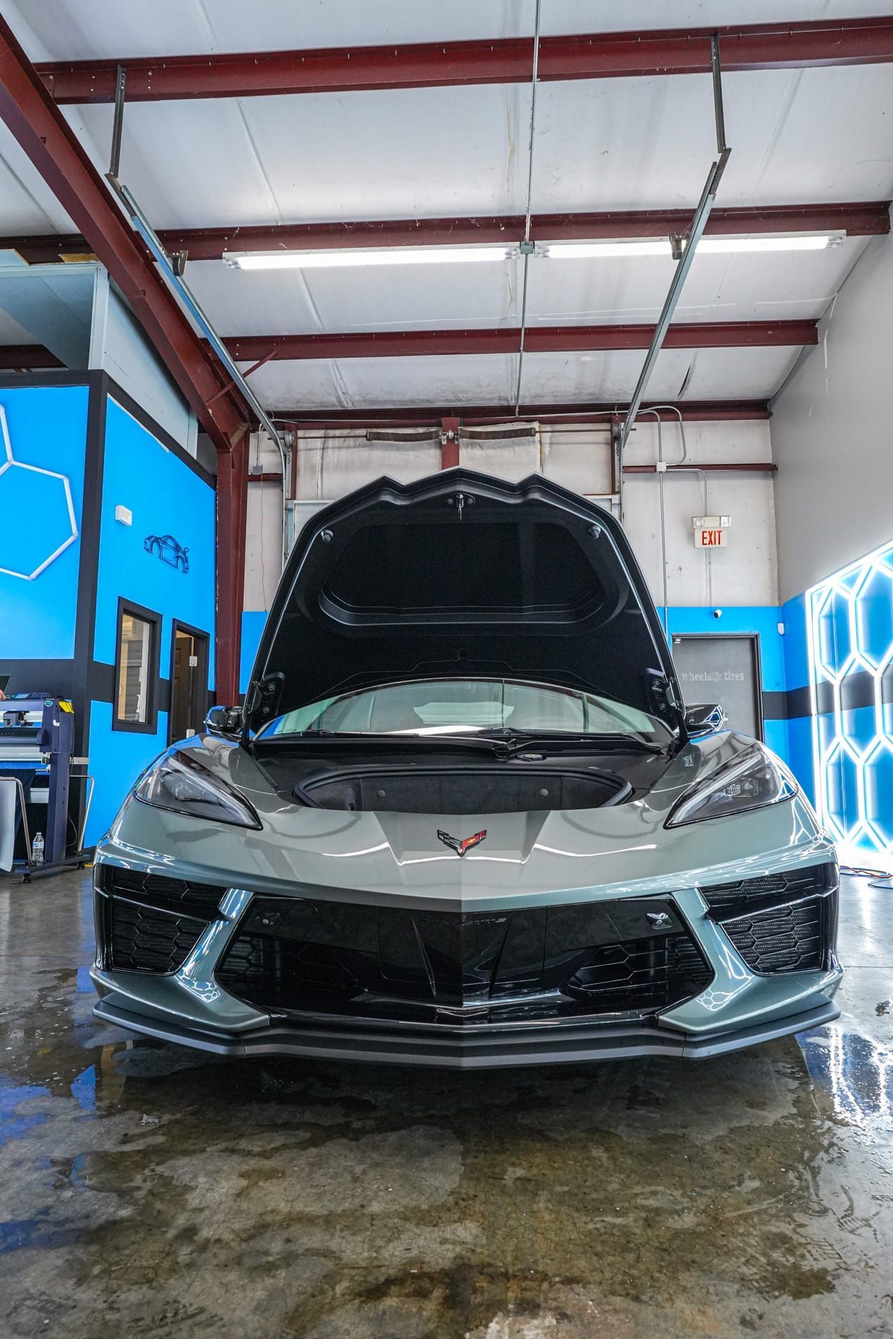 Gray sports car with hood open in a garage, blue accents, wet floor.