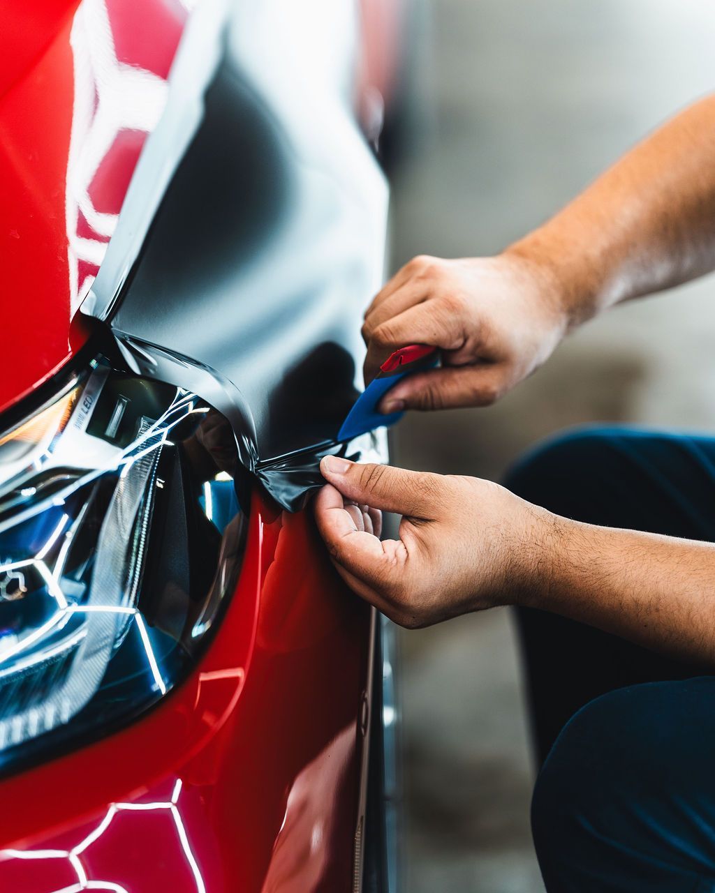 Person applying black vinyl wrap to a red car's bumper using a squeegee.