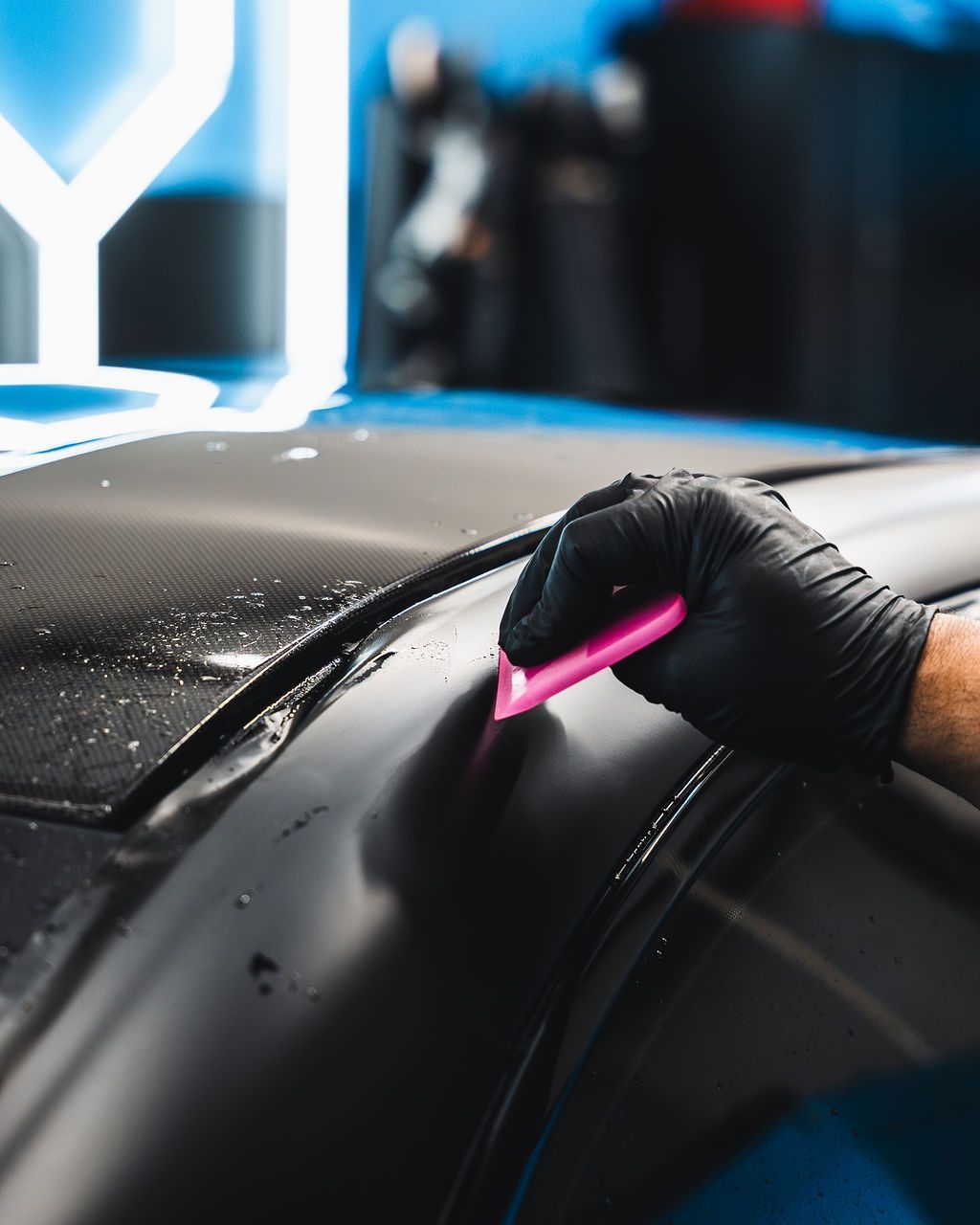 Gloved hand using a pink squeegee on a dark car surface, likely applying film.