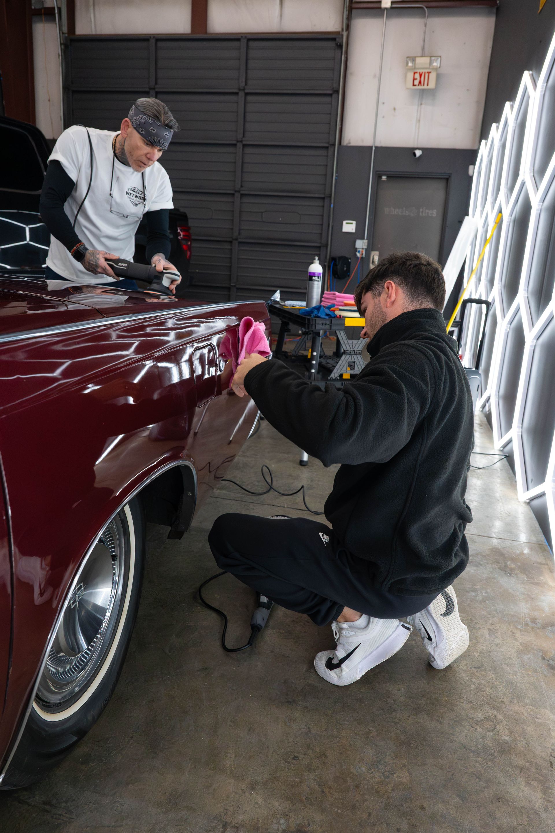 Two people polishing a maroon car in a garage with bright lights.