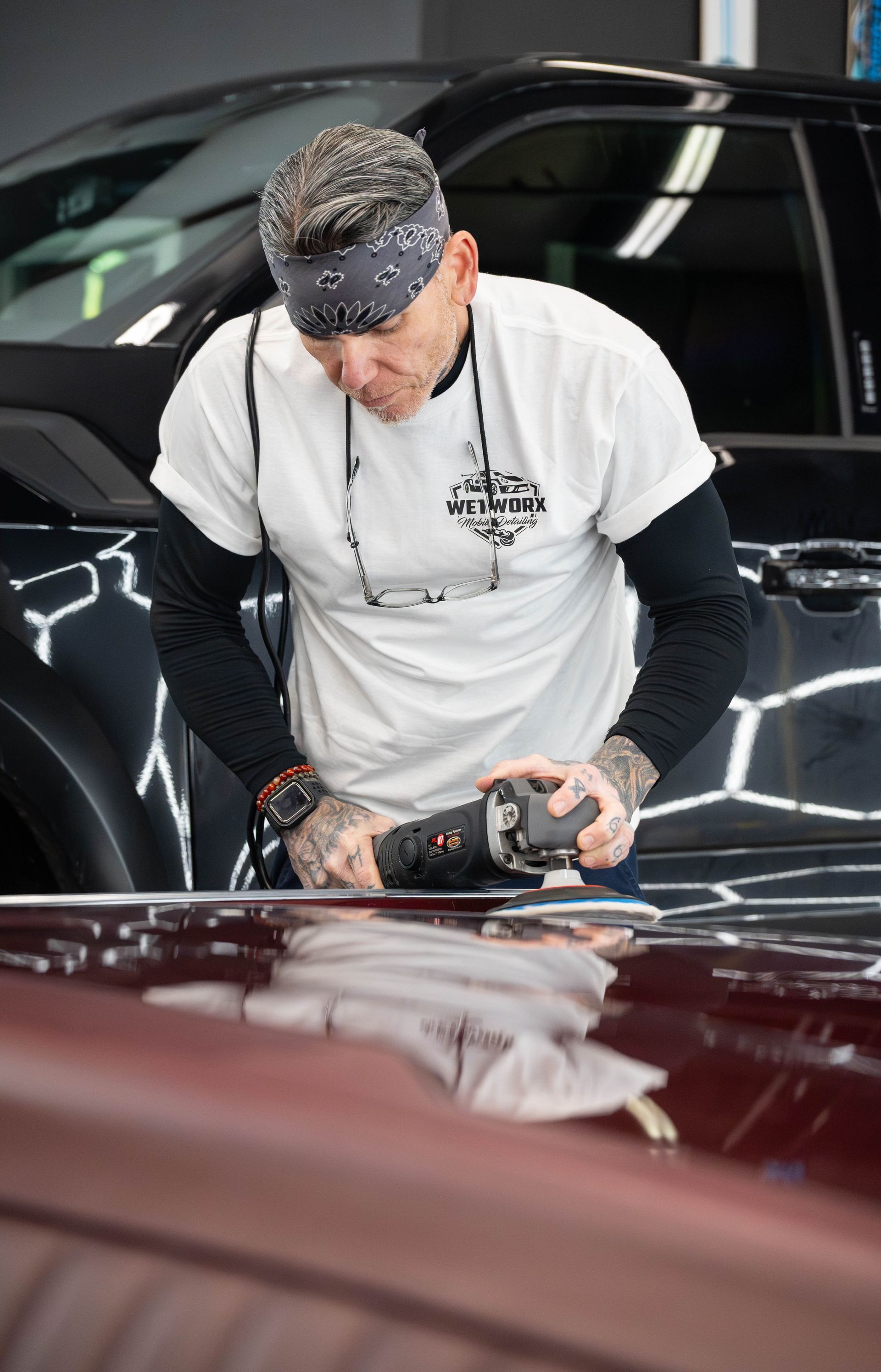 Man using a polisher on a car's hood in a garage. He wears a bandana, white t-shirt, and has tattoos on his arms.