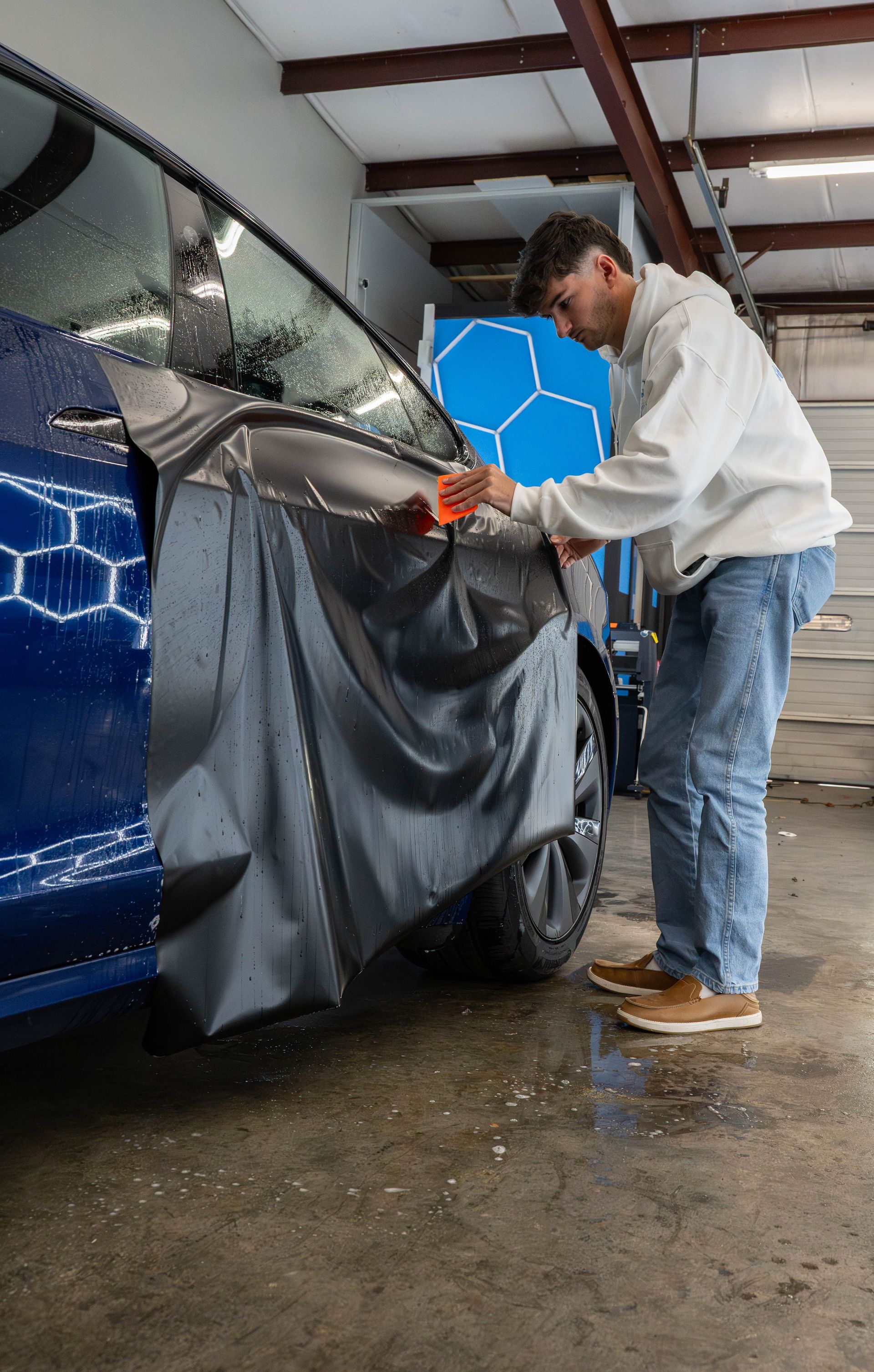 Man applying tinted film to a blue car window inside a garage.