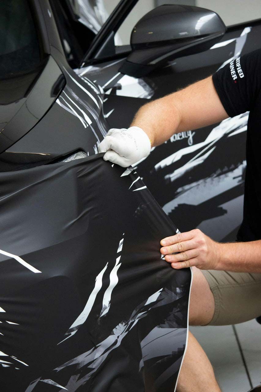 A person in gloves wrapping a dark patterned film onto a gray car panel.