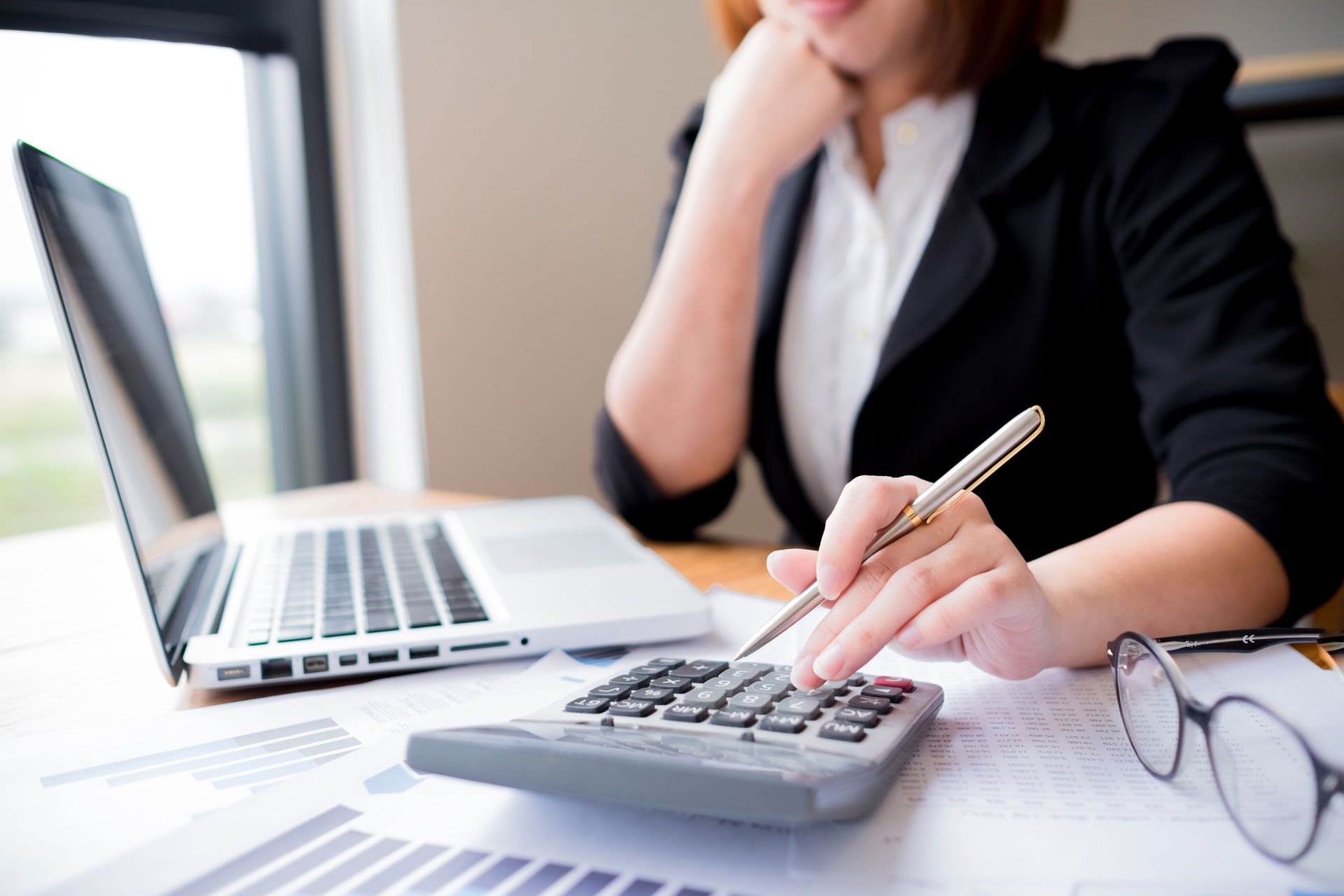 Woman in a black blazer calculating with a calculator, laptop, and glasses on a desk near a window.