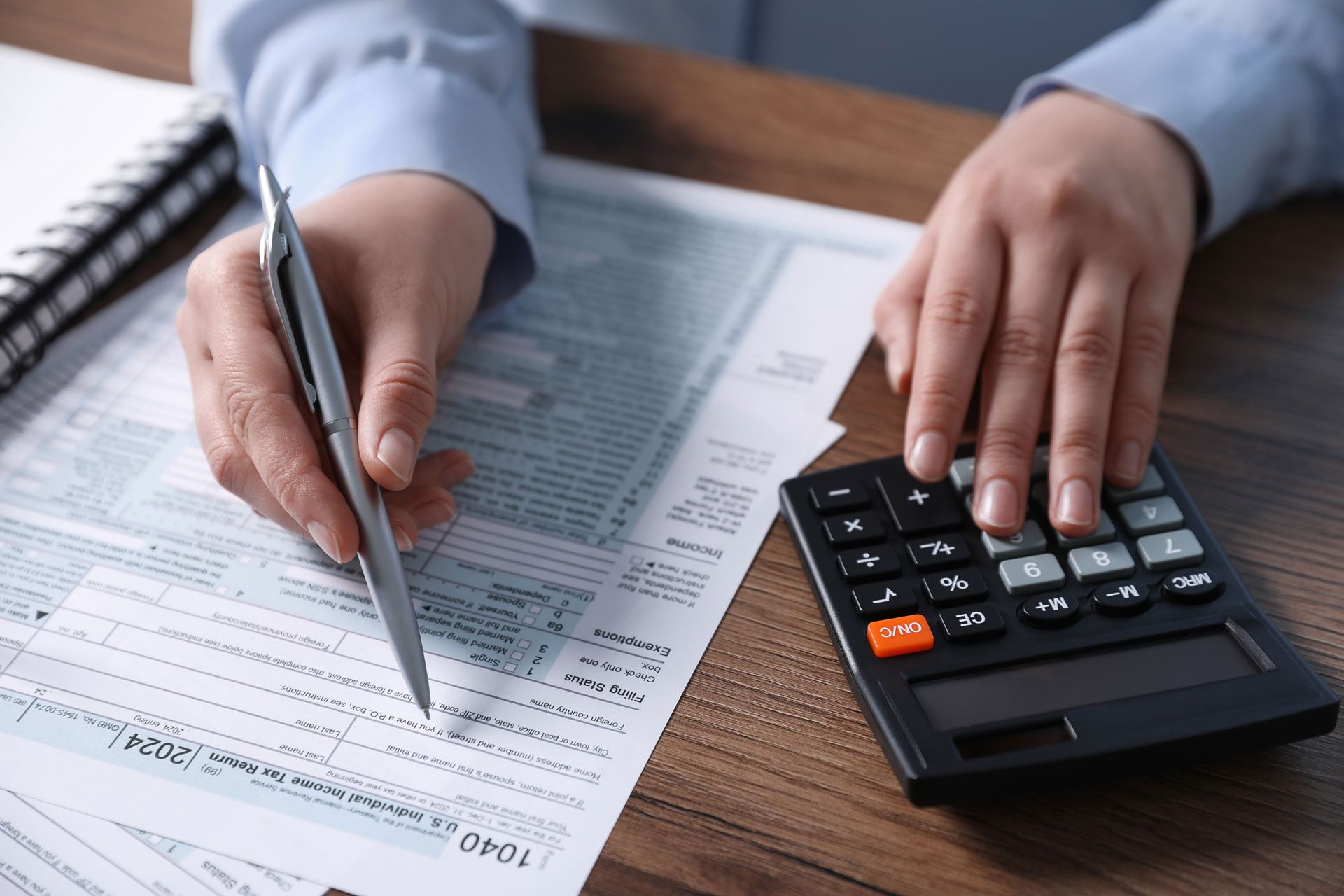 Person using a calculator and pen to fill out tax forms on a desk.