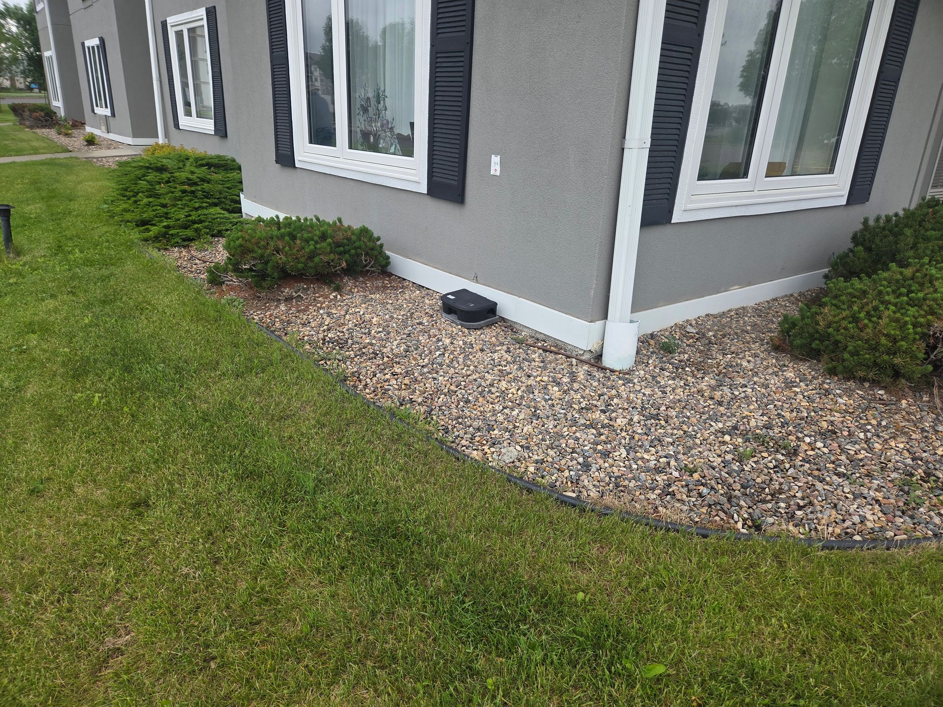 Gray house corner with white trim, gravel border, and green lawn in front.