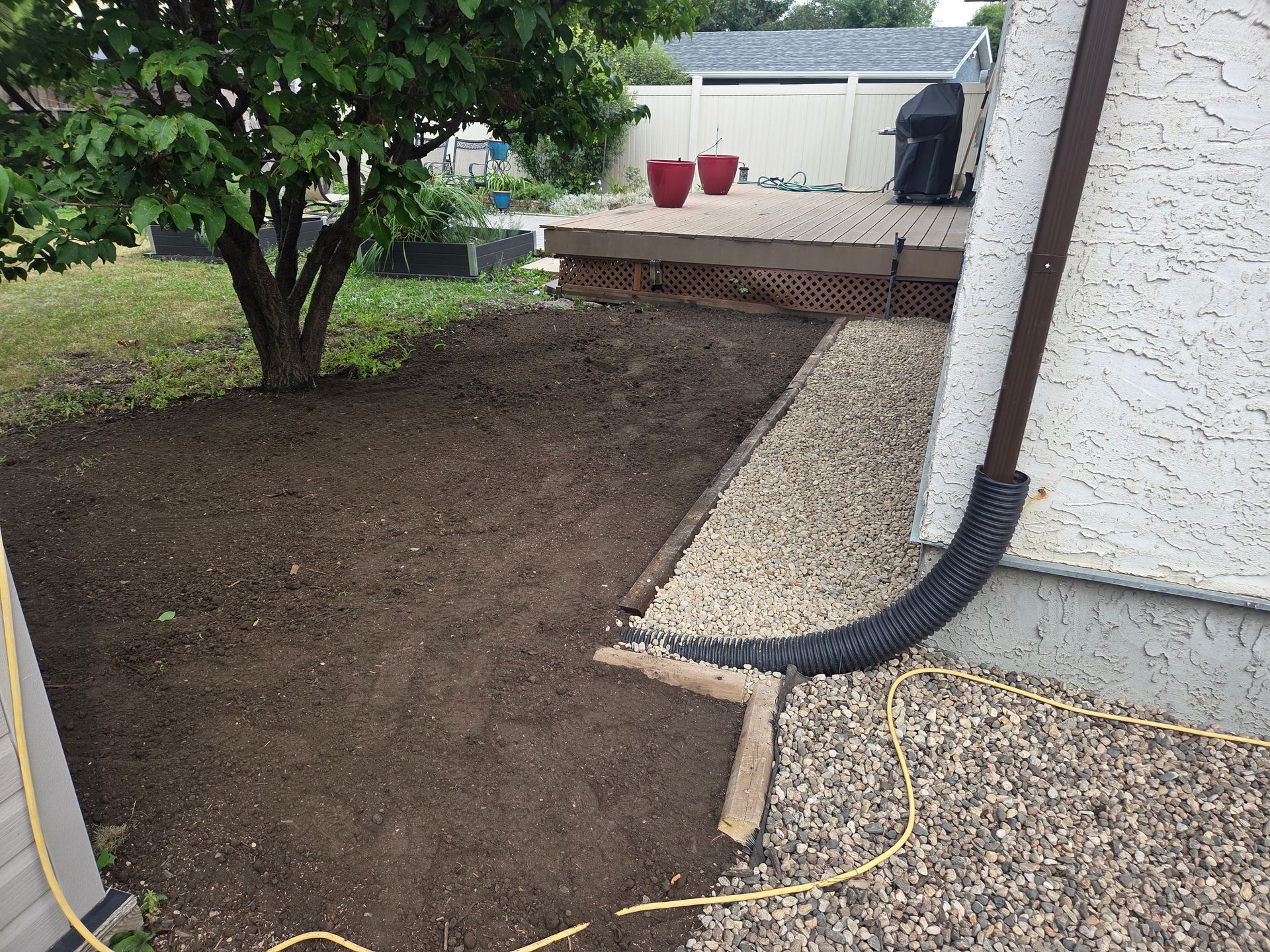 Freshly landscaped yard with dark soil, a tree, and gravel border beside a house wall and downspout.