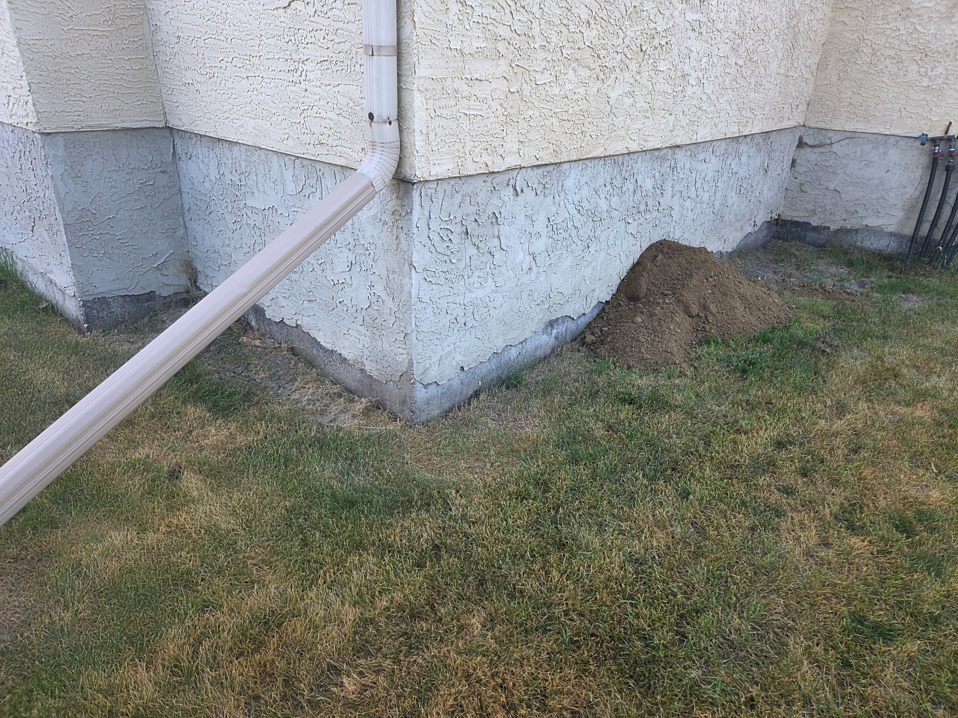 Building corner with drainage pipe and a small dirt mound beside a grassy lawn
