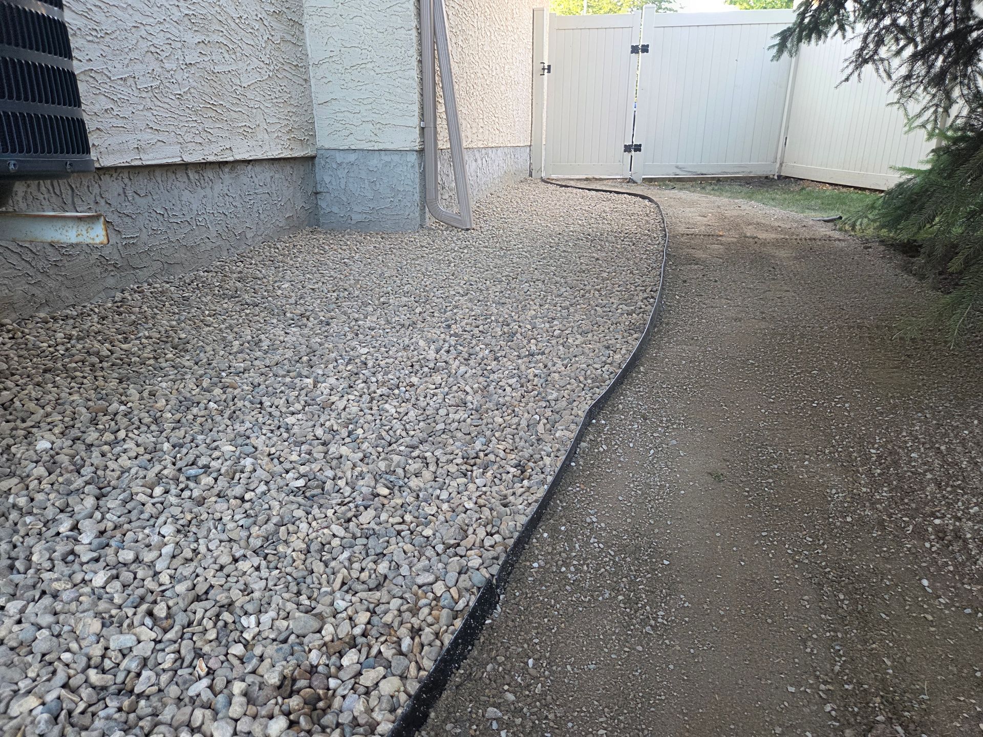 Gravel driveway beside a stucco house, curving toward a white gate and shrubbery.