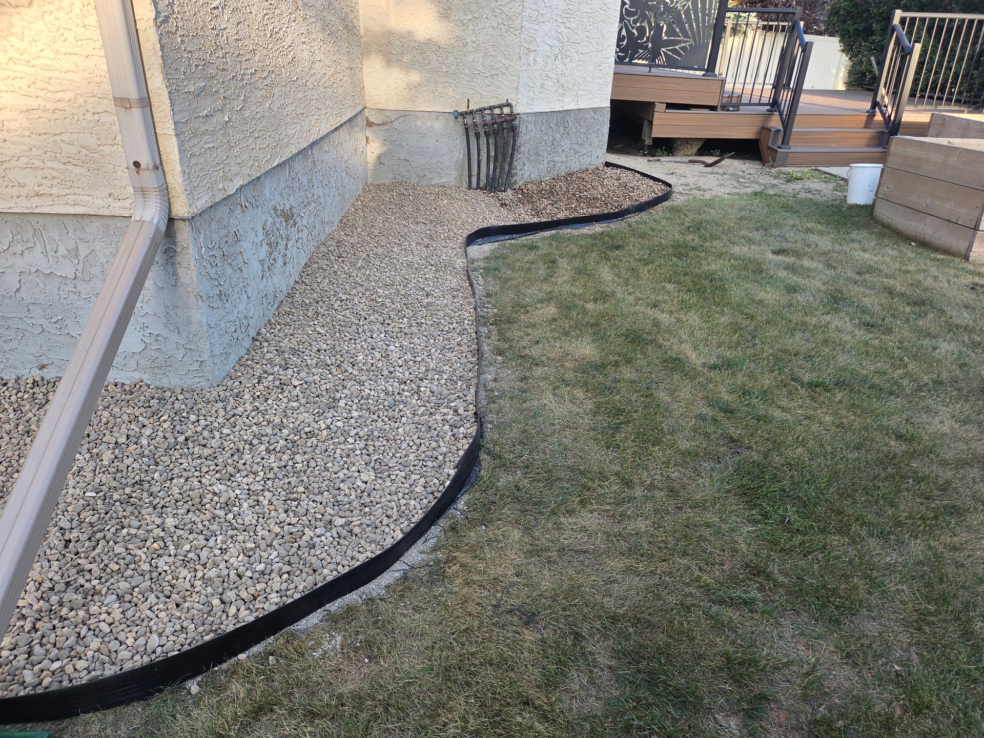 Curved gravel border along a house foundation beside a patchy lawn and small wooden deck area
