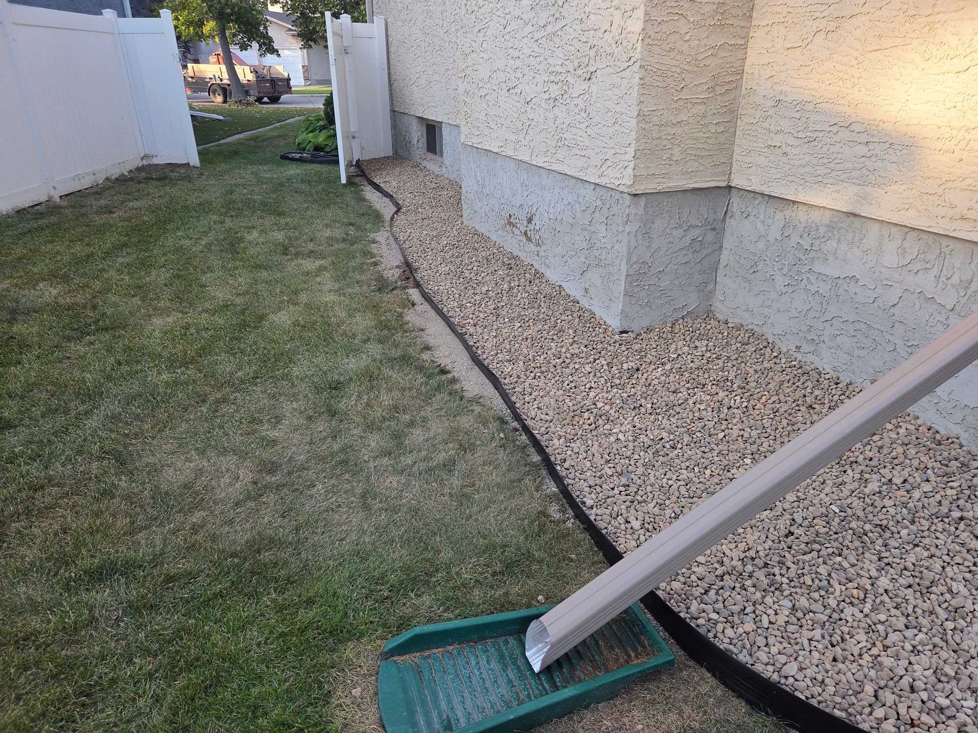 Lawn edge beside a house with a gravel strip and a green dustpan holding a long-handled tool