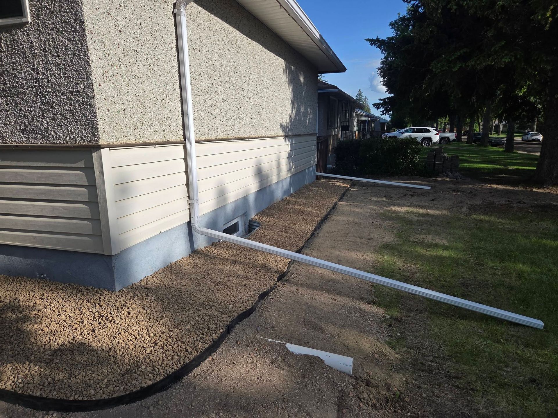 Side of a beige building with white trim, gravel strip, and a white pipe along a sidewalk under trees.