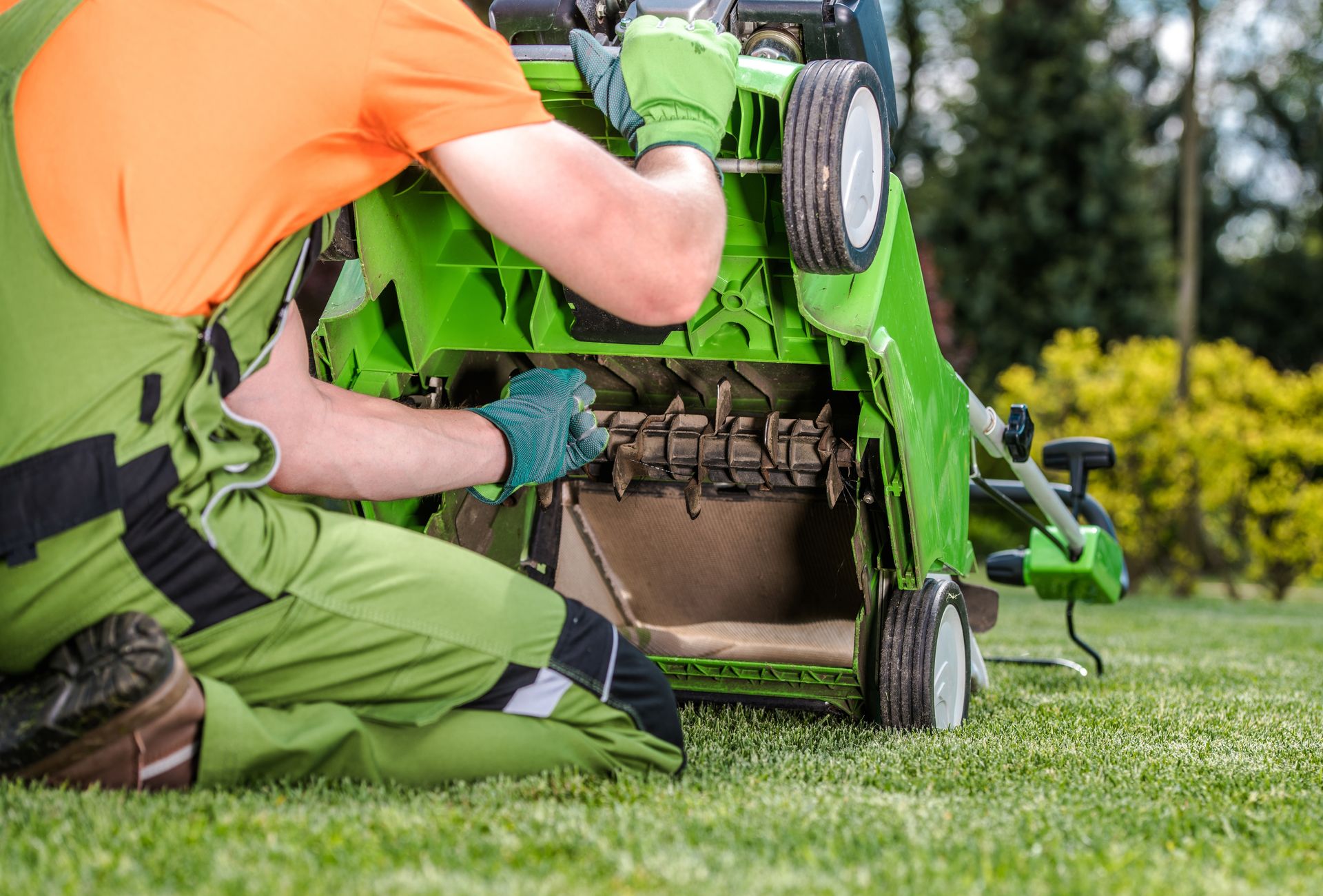 Worker repairing a green lawn mower on grass, wearing an orange safety vest and gloves
