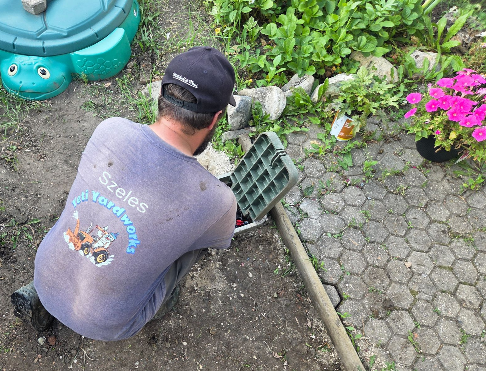 Person kneeling in a garden, digging soil beside pink flowers and a green edging tool.
