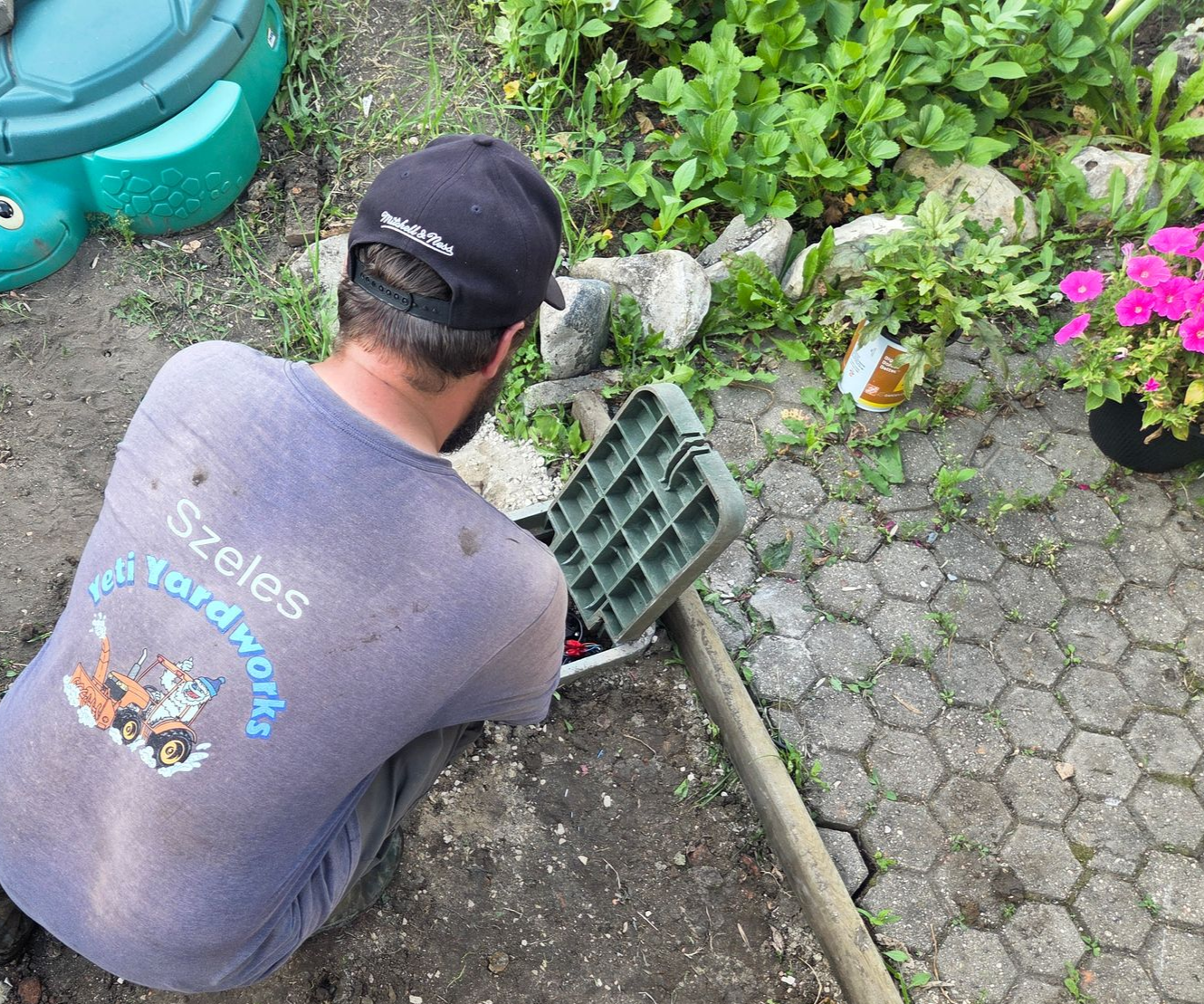 Person kneels in a garden, lifting a green crate beside flowers and paving stones.