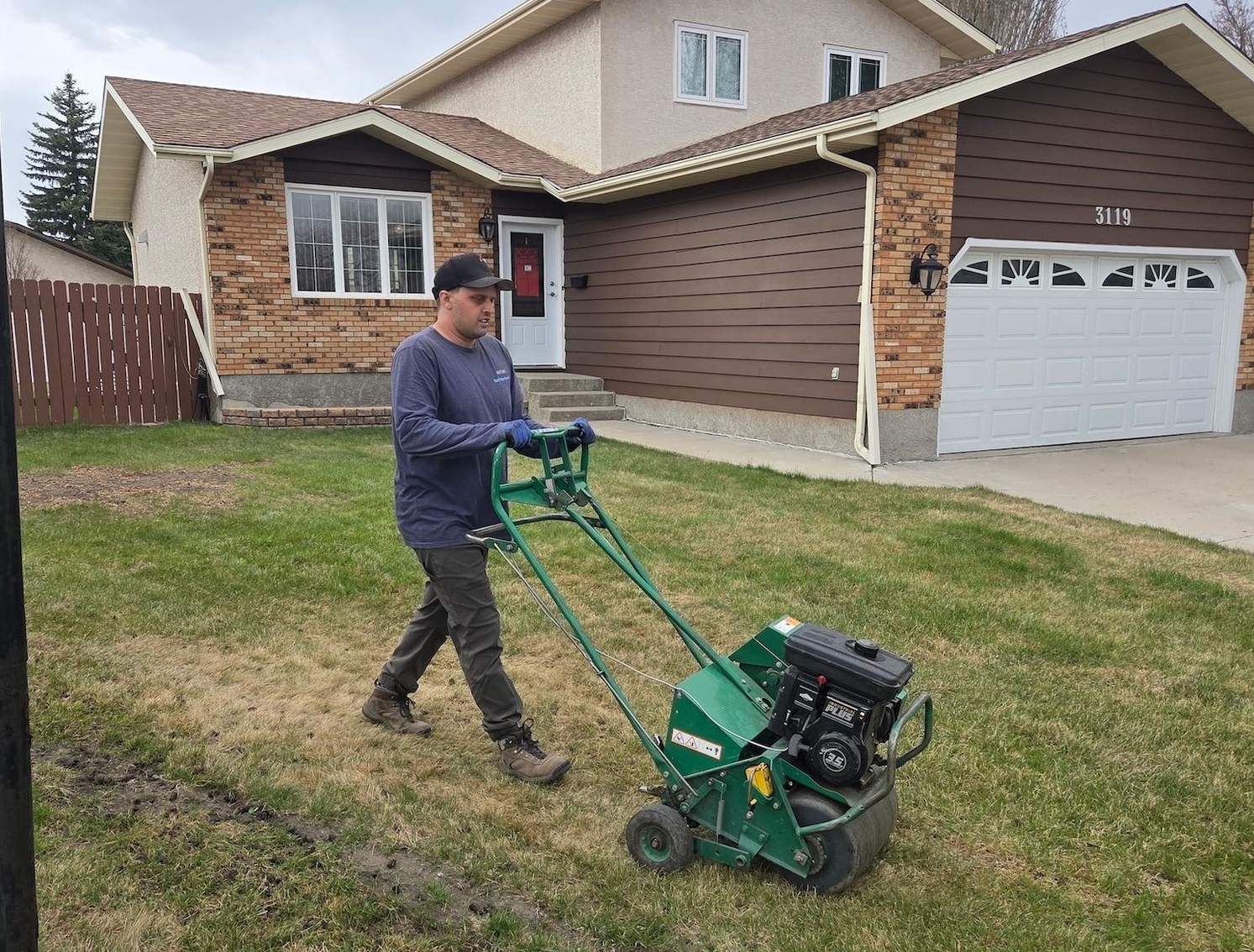 Person using a green lawn aerator on a suburban front yard near a house and garage.