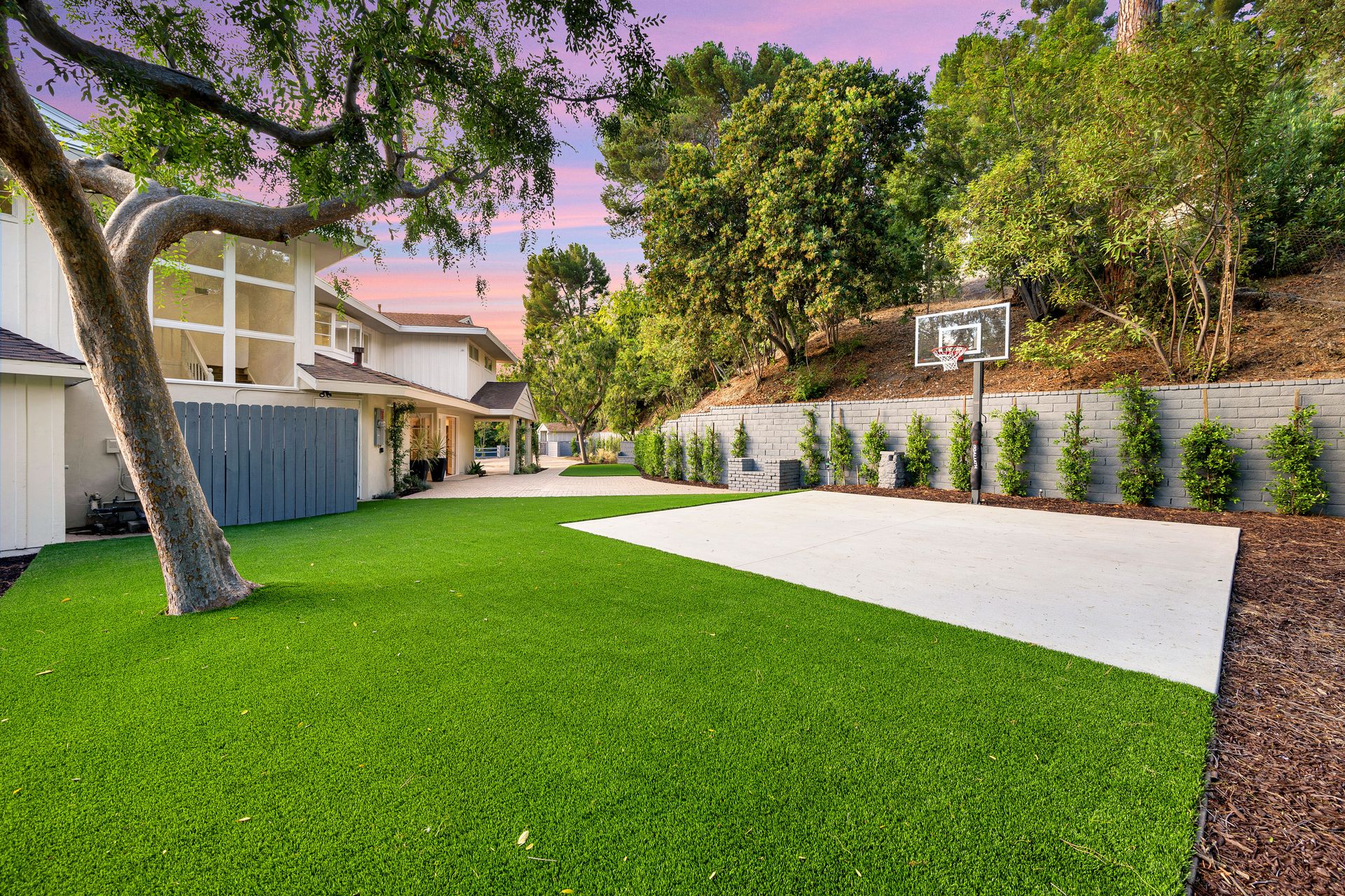 Backyard with lush green lawn, white patio, tree, and basketball hoop beside a fence