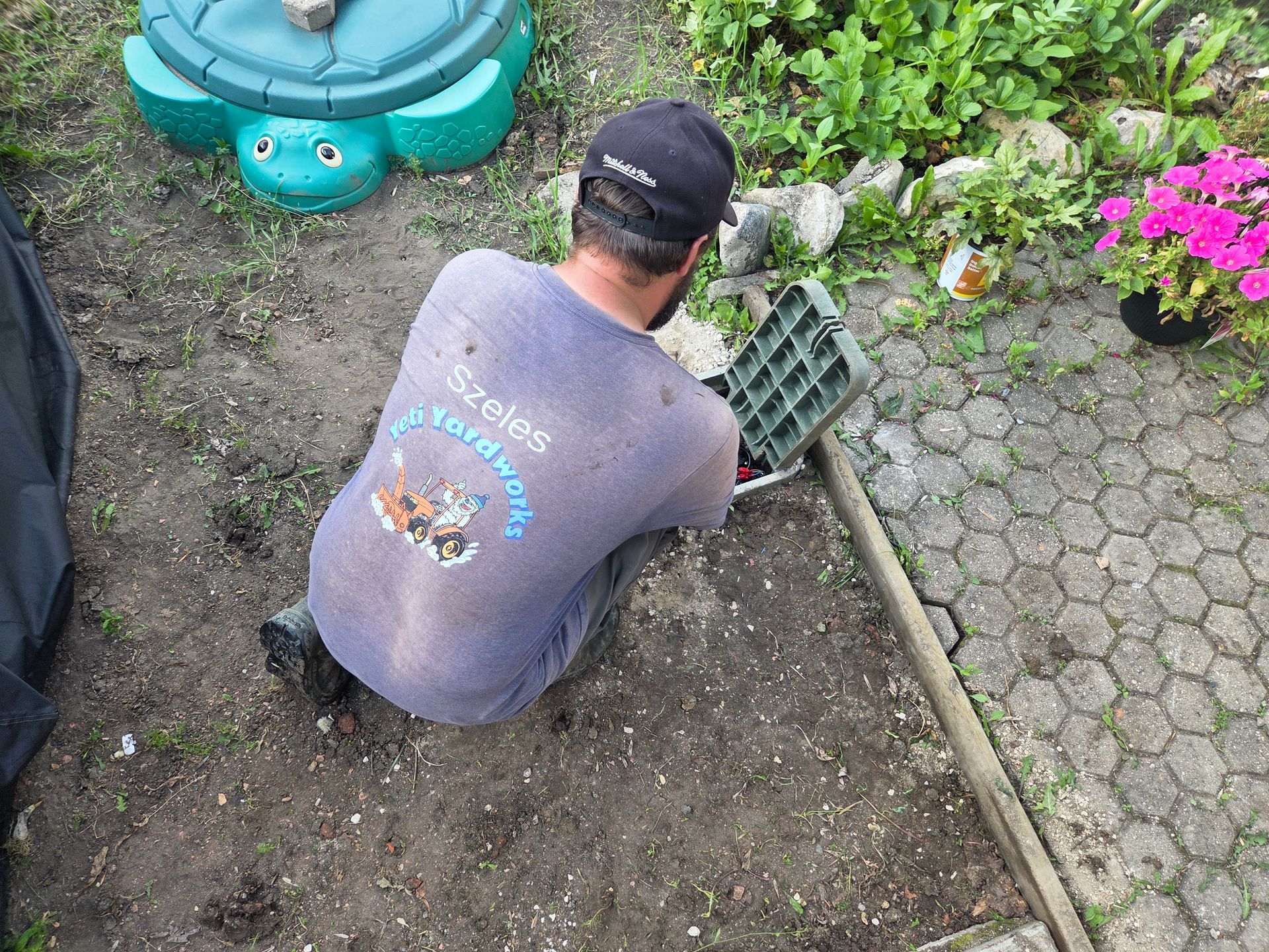 Person kneeling in a garden, filling soil beside a paved path and flower bed