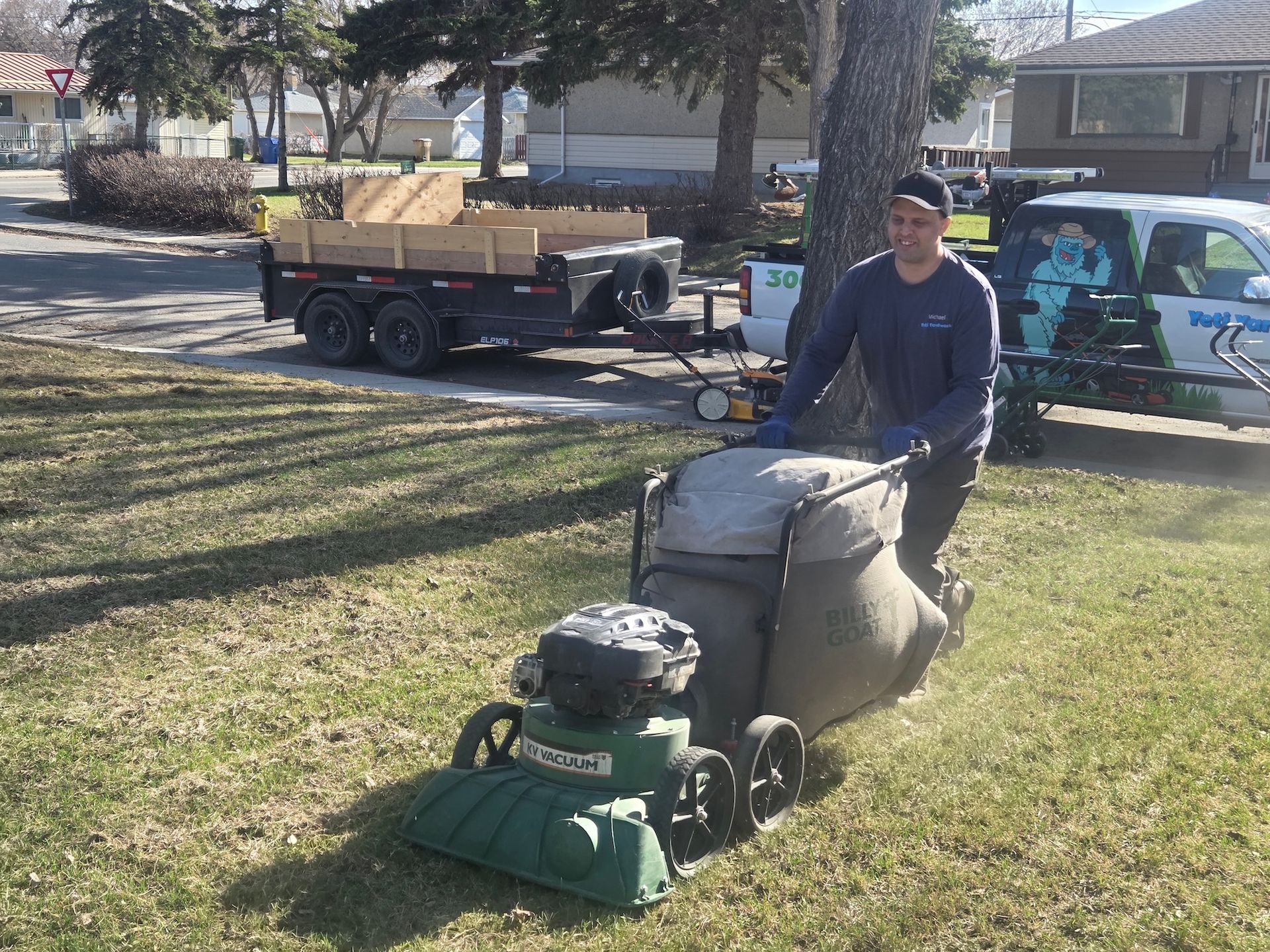 Person pushing a green lawn mower on a grassy park lawn beside a trailer and parked car.