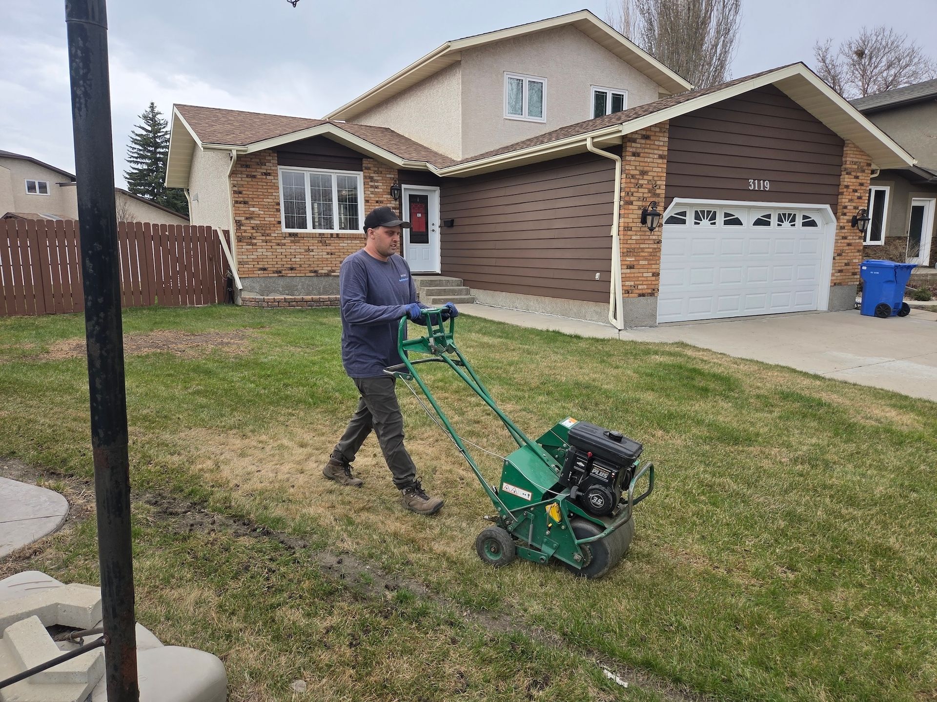 Person mowing a suburban lawn with a green push mower in front of a brick house.