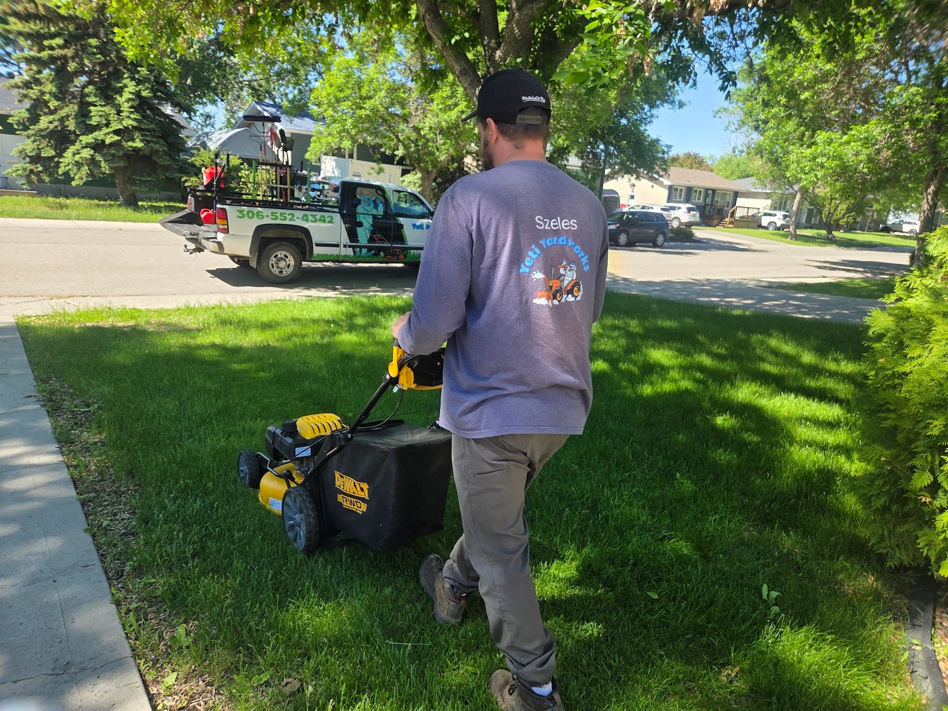 Person mowing a lawn with a push mower beside a sidewalk in a sunny neighborhood.