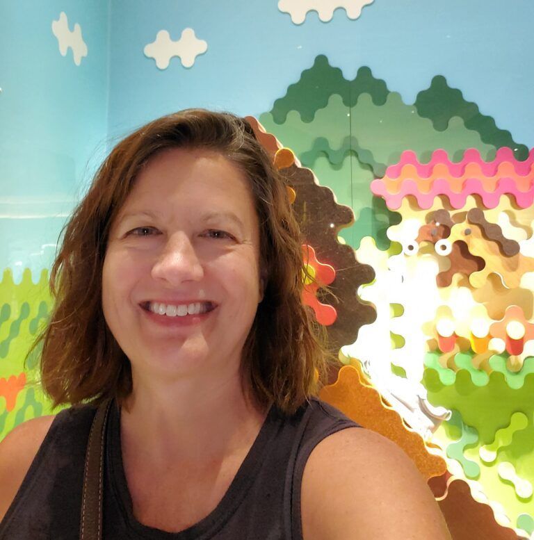 Woman smiling, posing in front of colorful wall art depicting hills, clouds, and animal.