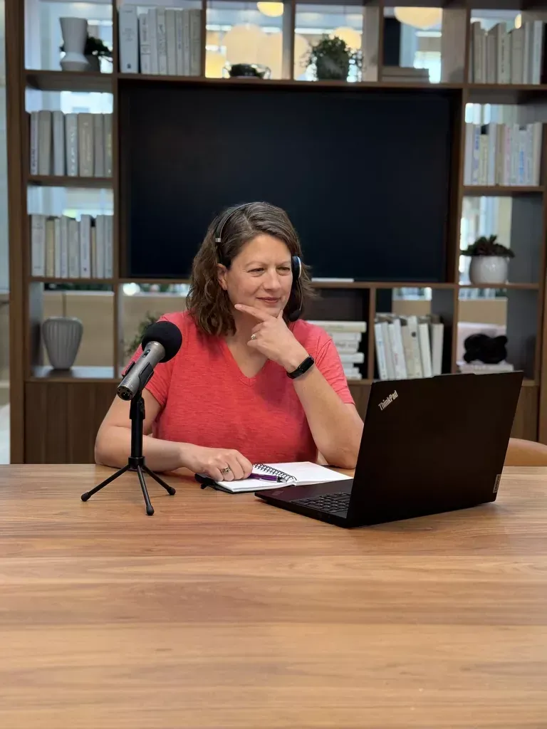 Woman seated at a table speaking into a microphone beside a laptop in a studio setup