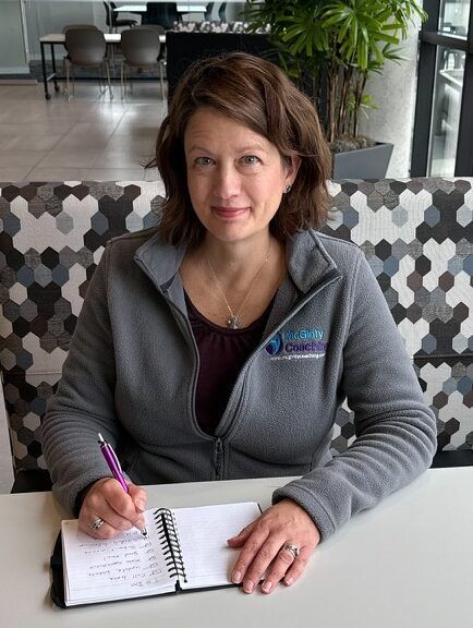 Woman in a gray jacket sitting at a table, writing in a notebook, smiling.