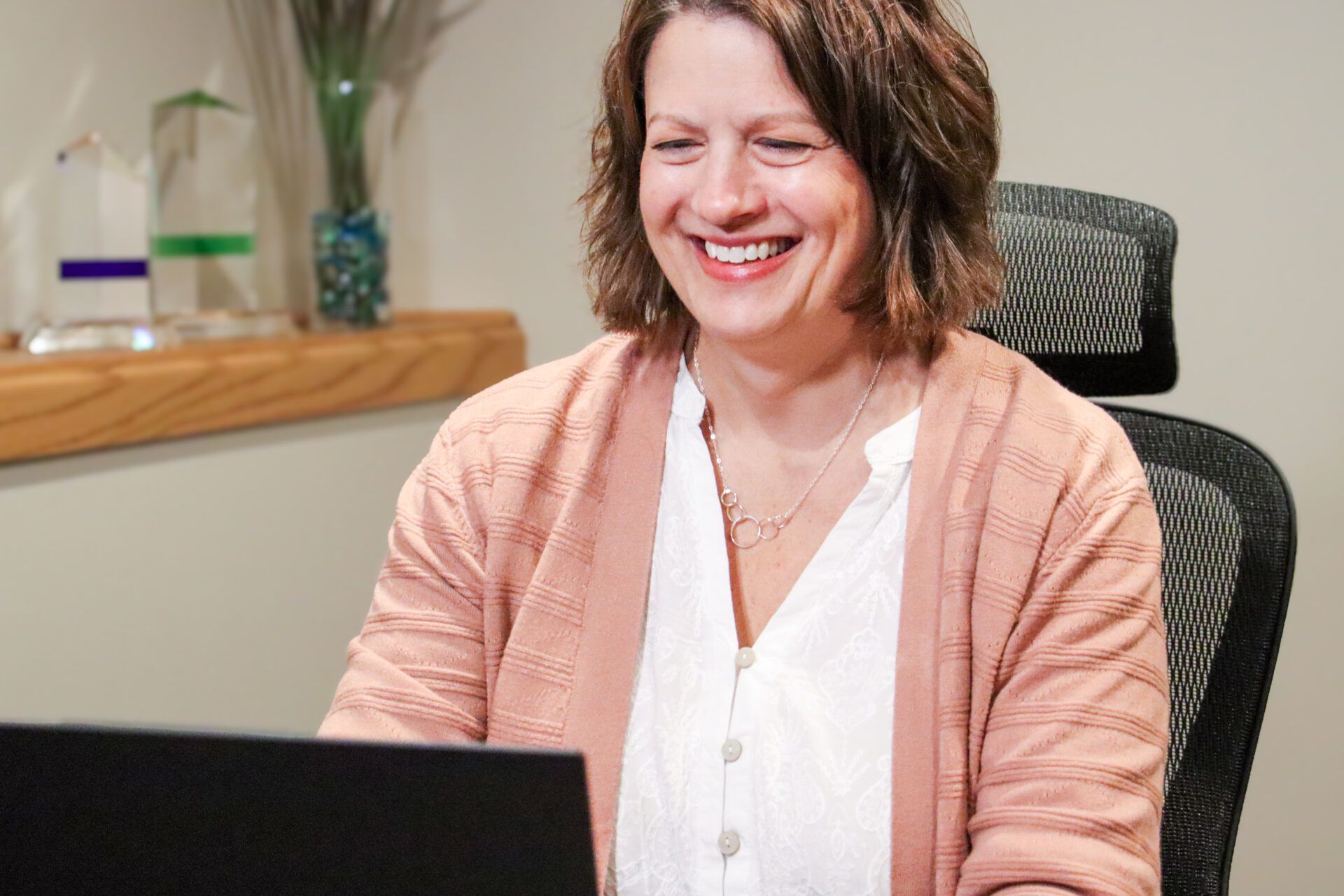 Smiling woman in a pink cardigan using a laptop at a desk in an office
