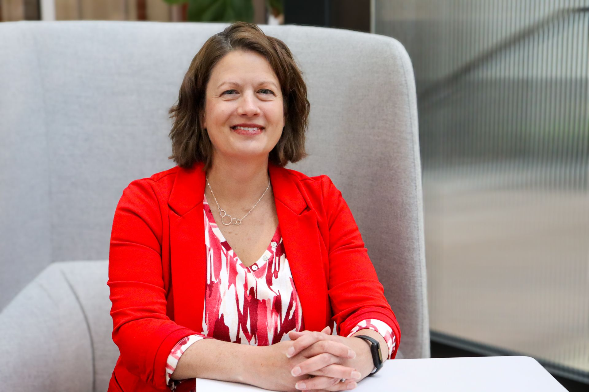 Woman in a red blazer sitting in a chair, smiling with hands clasped at a table.