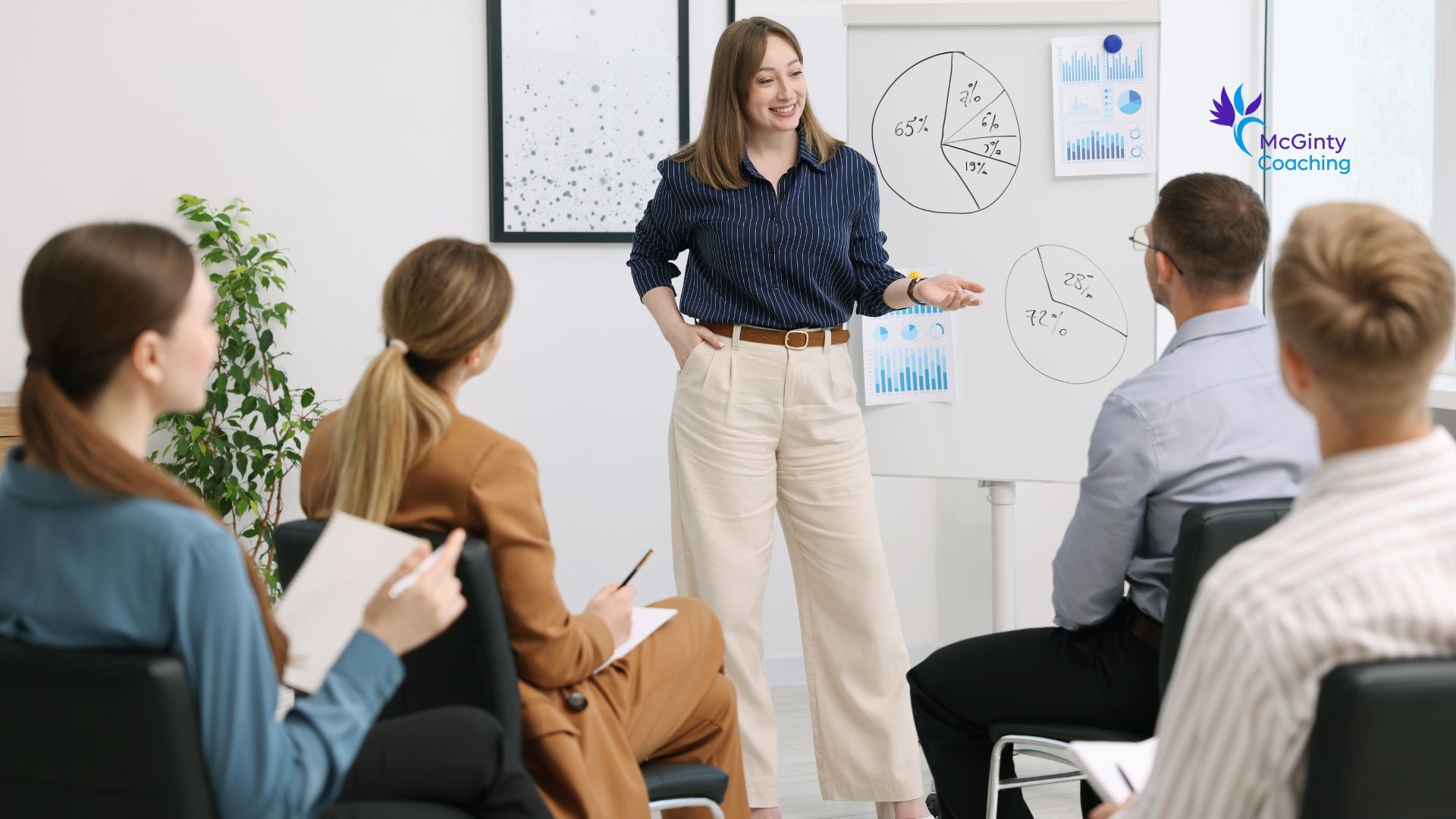 Presenter speaking to a seated group in a bright meeting room with a whiteboard and charts.